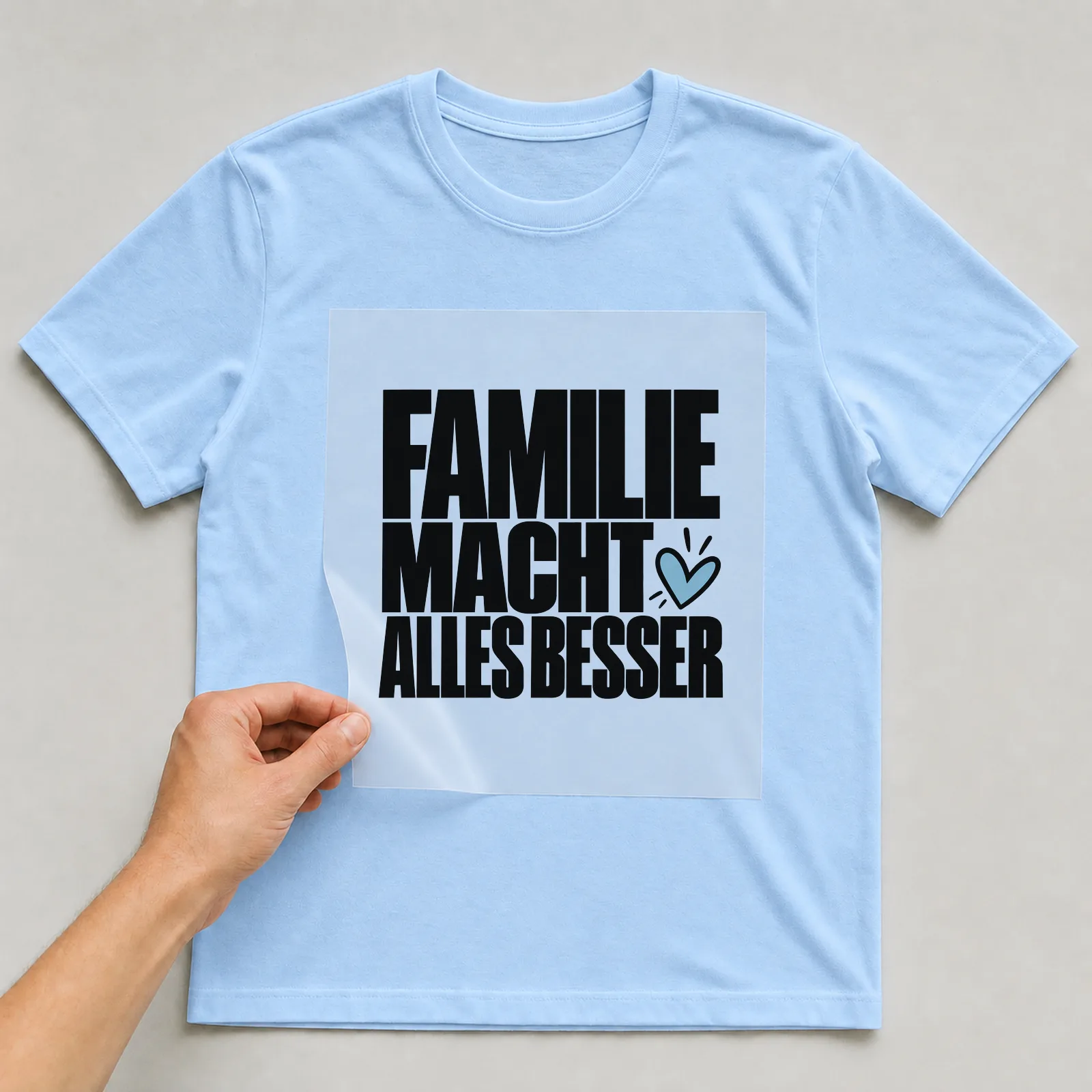 Bügelbild „Familie macht alles besser“ auf hellblauem T-Shirt, Folie wird aufgelegt