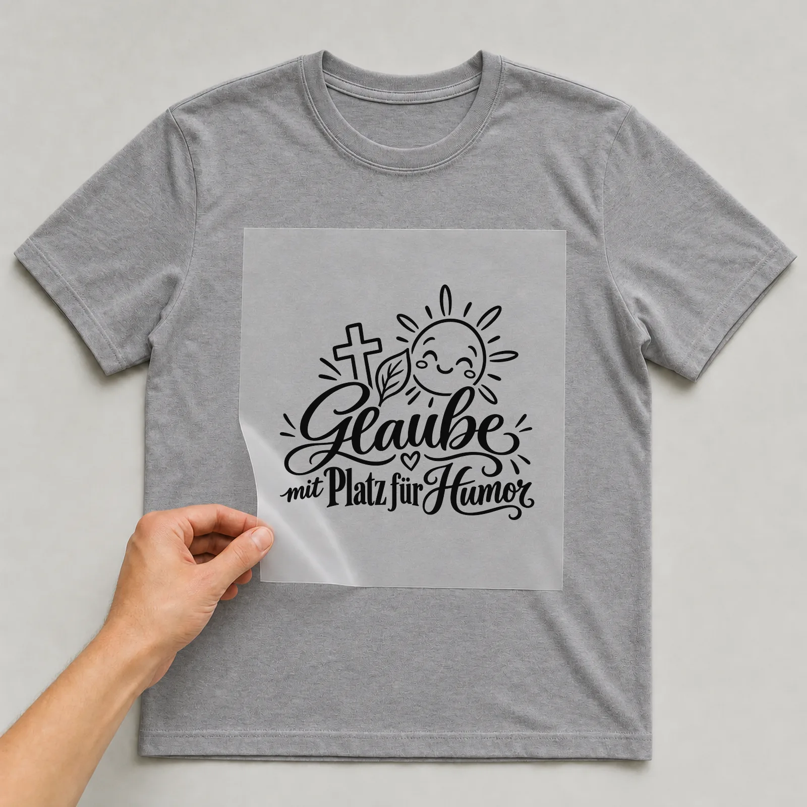 Bügelbild „Glaube mit Platz für Humor“ auf grauem T-Shirt, Folie wird aufgelegt