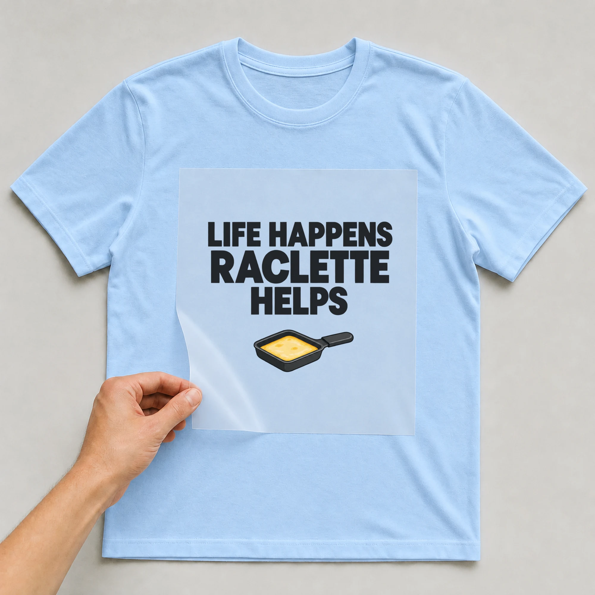 Bügelbild „LIFE HAPPENS RACLETTE HELPS“ auf babyblauem T-Shirt, Folie wird aufgelegt
