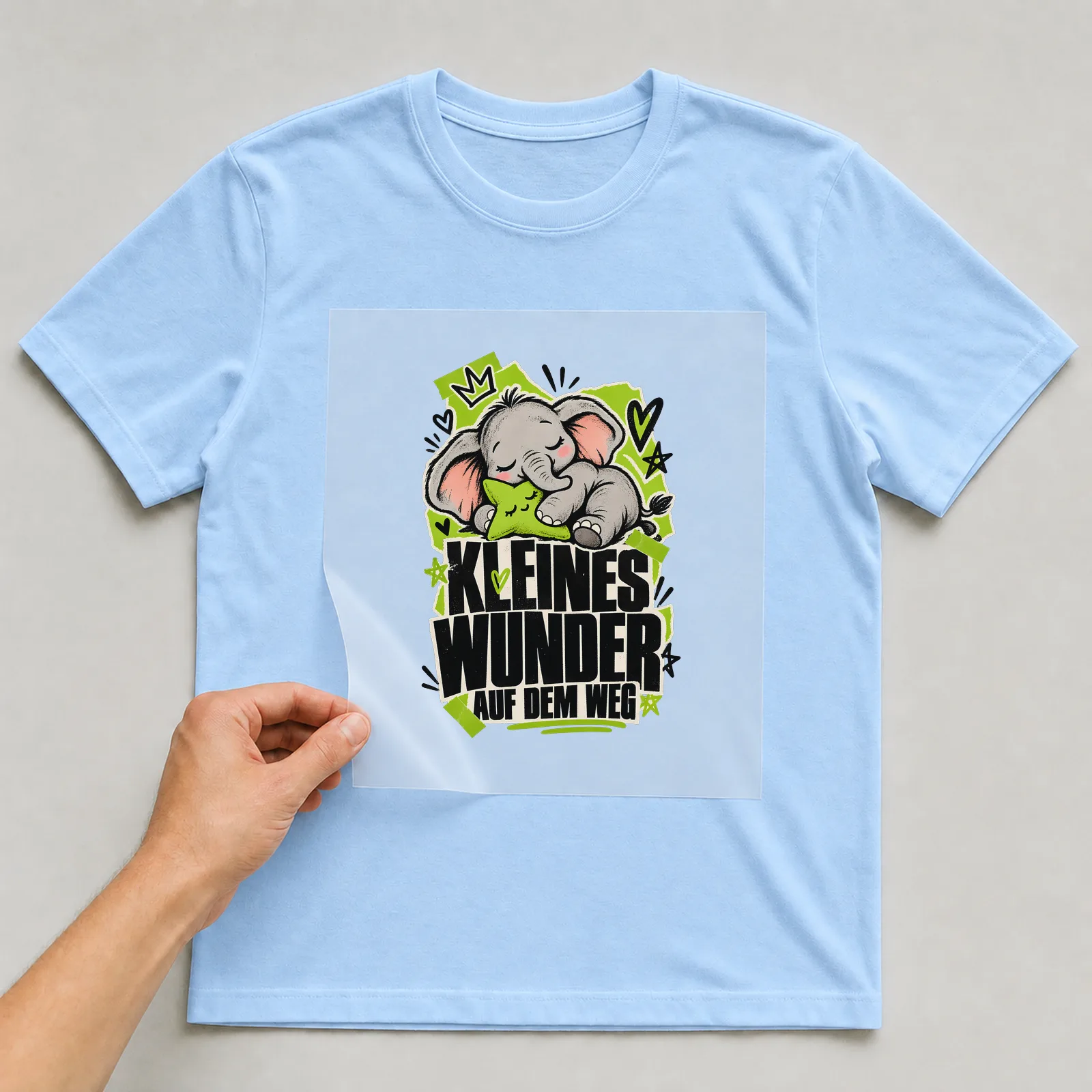 Bügelbild „Kleines Wunder auf dem Weg“ auf hellblauem T-Shirt, Folie wird aufgelegt