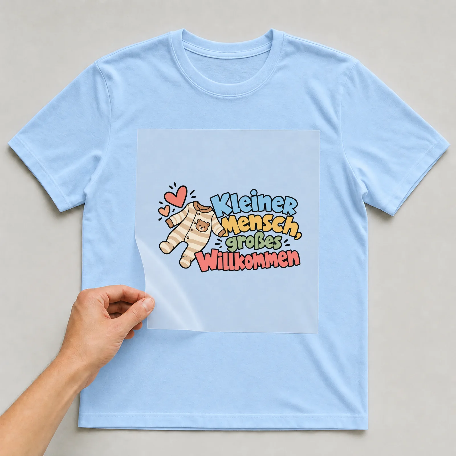 Bügelbild „Kleiner Mensch, großes Willkommen“ auf hellblauem T-Shirt, Folie wird aufgelegt