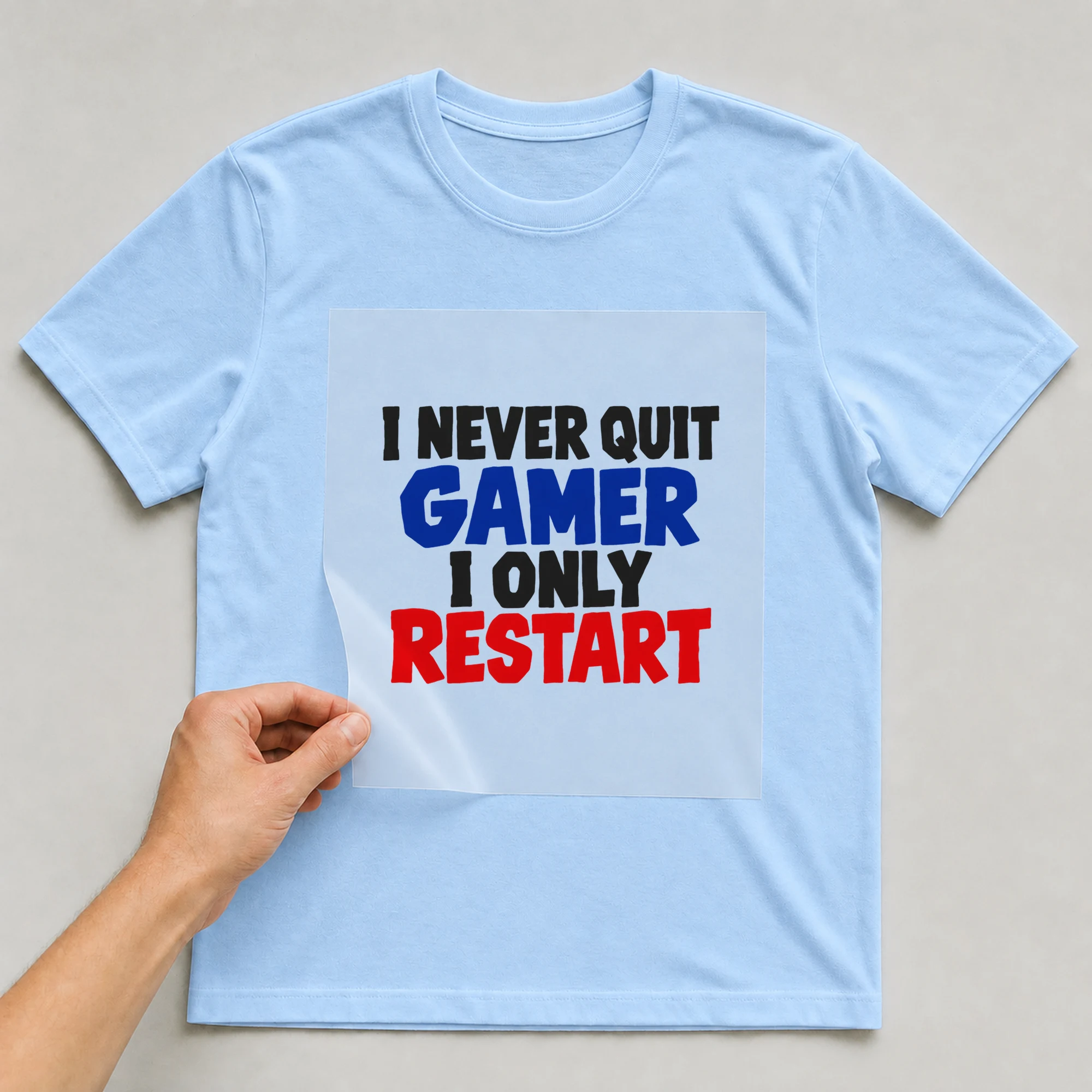 Bügelbild „I Never Quit Gamer I Only Restart“ auf babyblauem T-Shirt, Folie wird aufgelegt