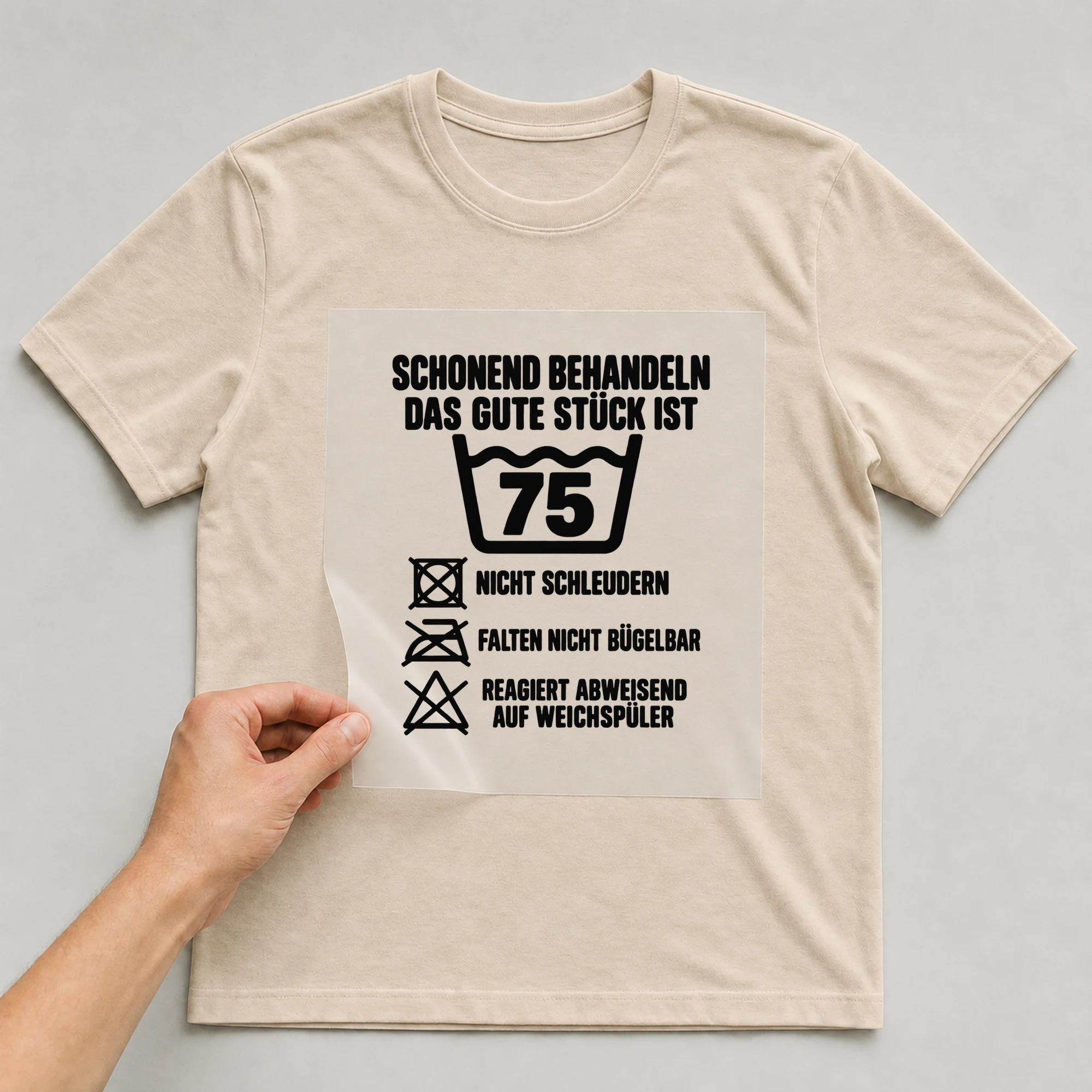 Bügelbild „Schonend behandeln das gute Stück ist 75 nicht schleudern falten nicht“ auf beigem T-Shirt, Folie wird aufgelegt