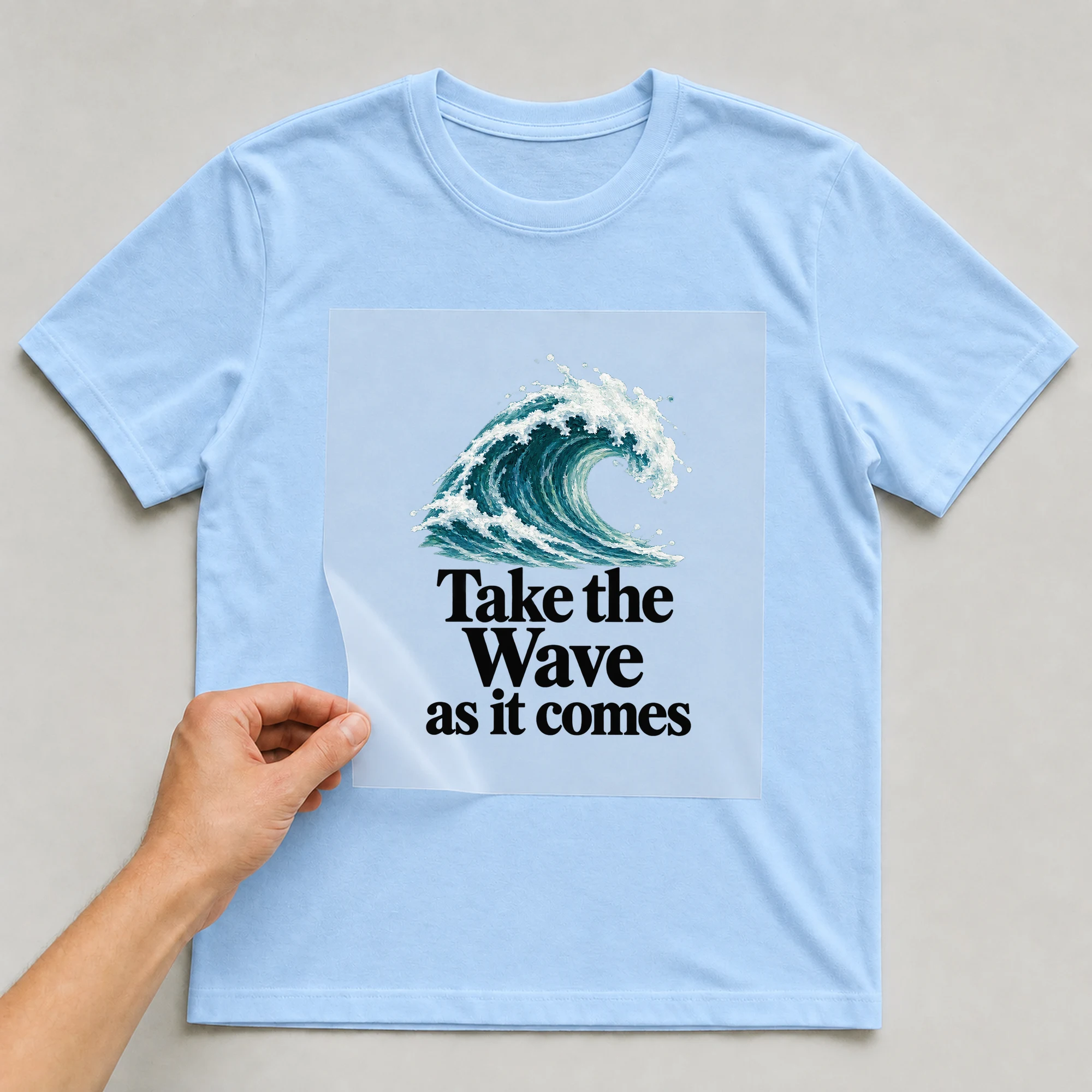 Bügelbild „Take the Wave as it comes“ auf babyblauem T-Shirt, Folie wird aufgelegt