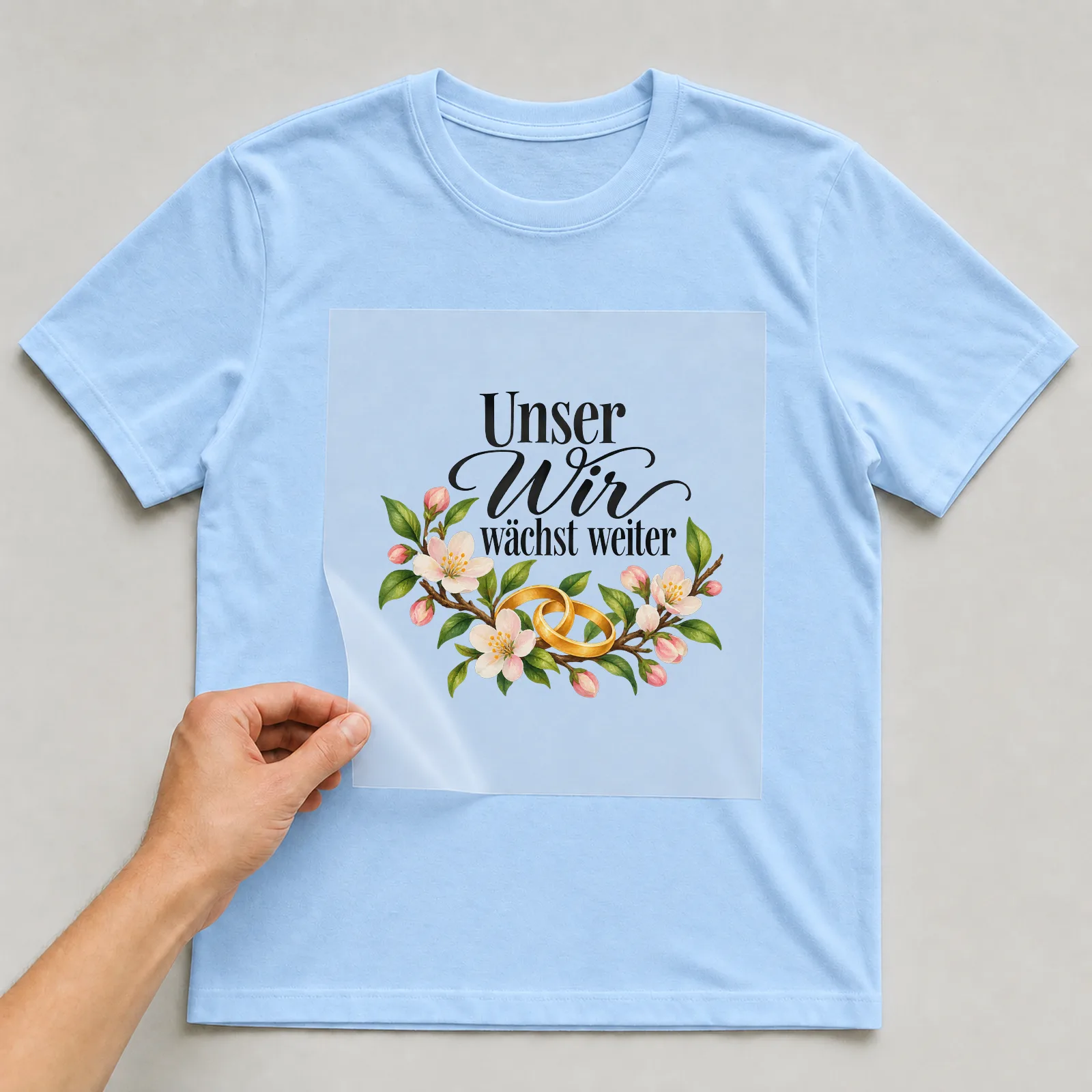Bügelbild „Unser Wir wächst weiter“ auf hellblauem T-Shirt, Folie wird aufgelegt