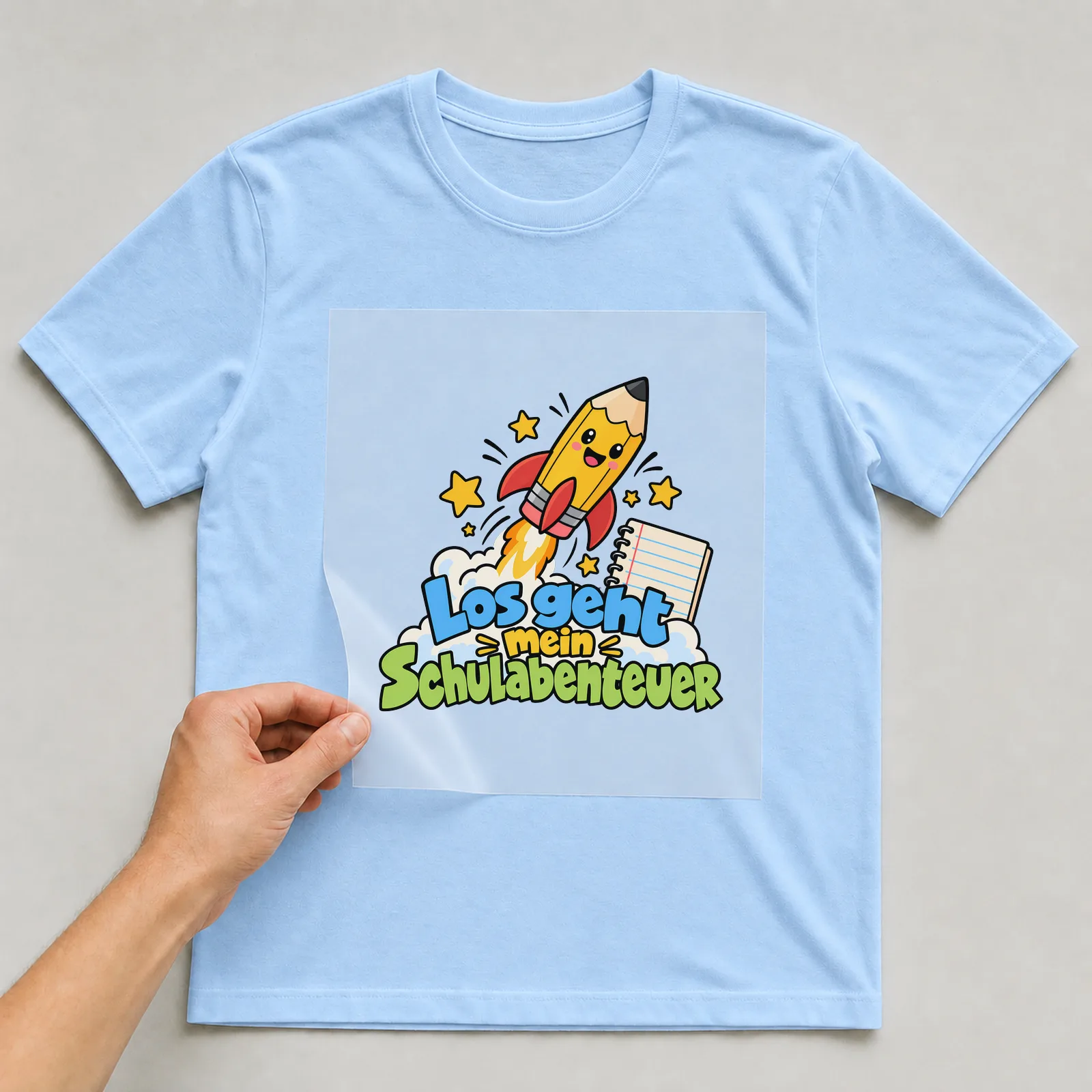 Bügelbild „Los geht mein Schulabenteuer“ auf hellblauem T-Shirt, Folie wird aufgelegt