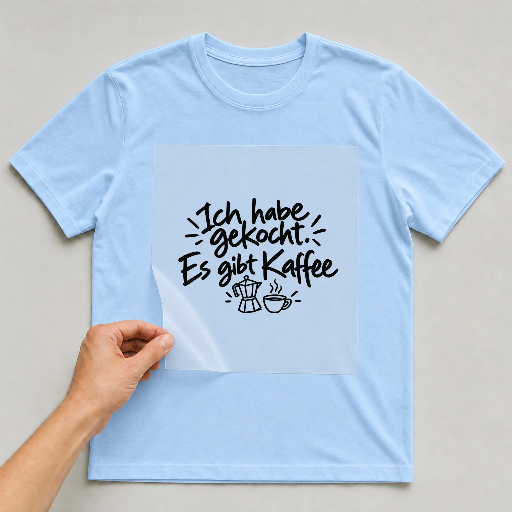 Bügelbild „Ich habe gekocht. Es gibt Kaffee“ auf babyblauem T-Shirt, Folie wird aufgelegt