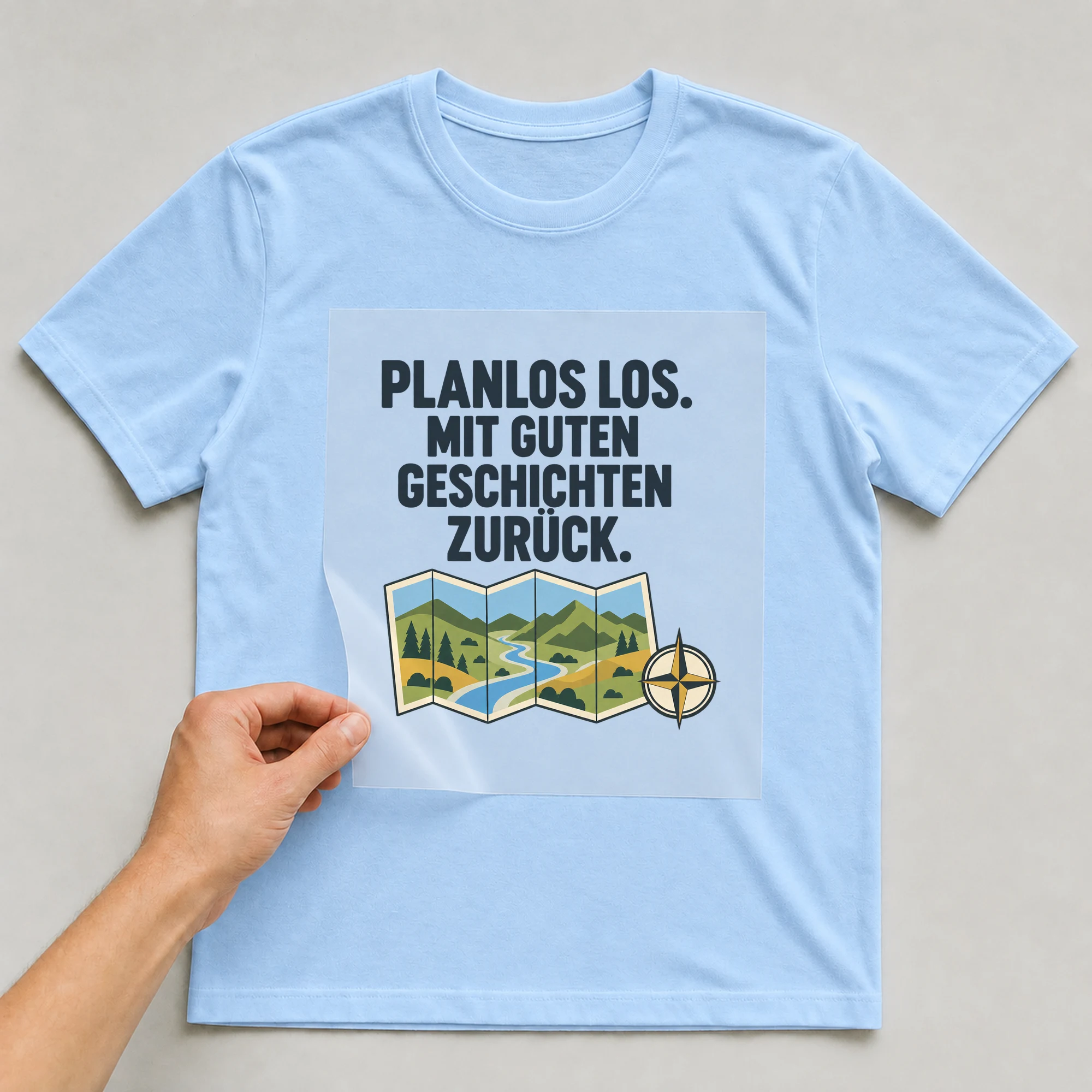 Bügelbild „PLANLOS LOS. MIT GUTEN GESCHICHTEN ZURÜCK.“ auf babyblauem T-Shirt, Folie wird aufgelegt