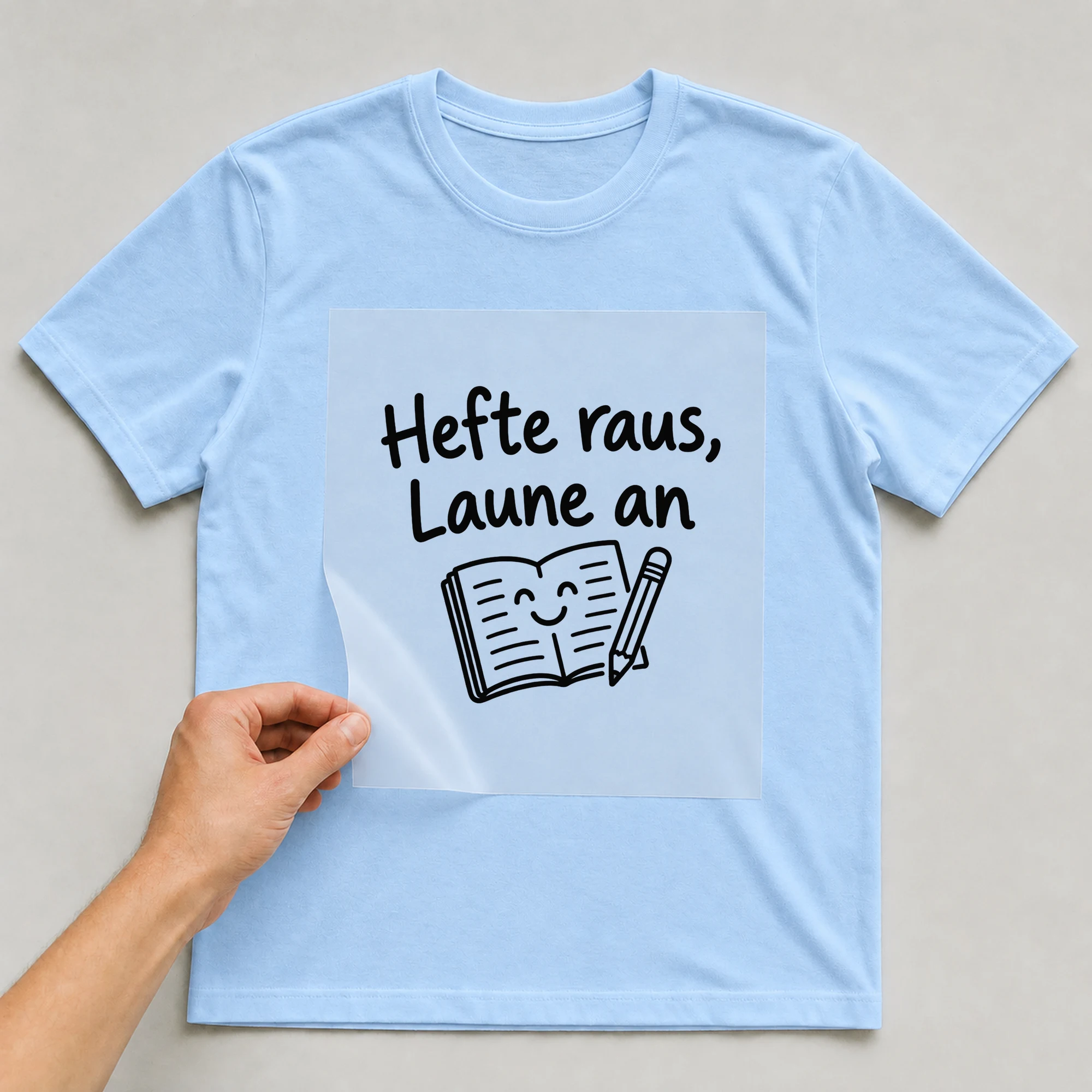 Bügelbild „Hefte raus, Laune an“ auf babyblauem T-Shirt, Folie wird aufgelegt