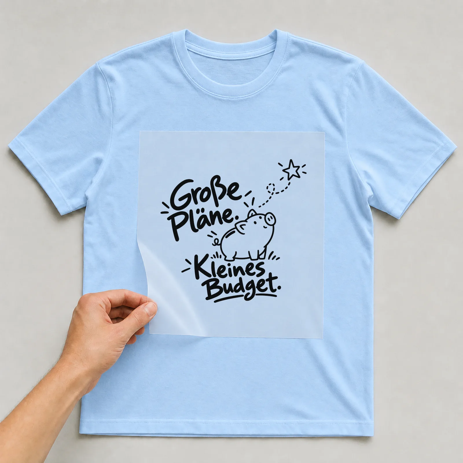 Bügelbild „Große Pläne. Kleines Budget.“ auf hellblauem T-Shirt, Folie wird aufgelegt
