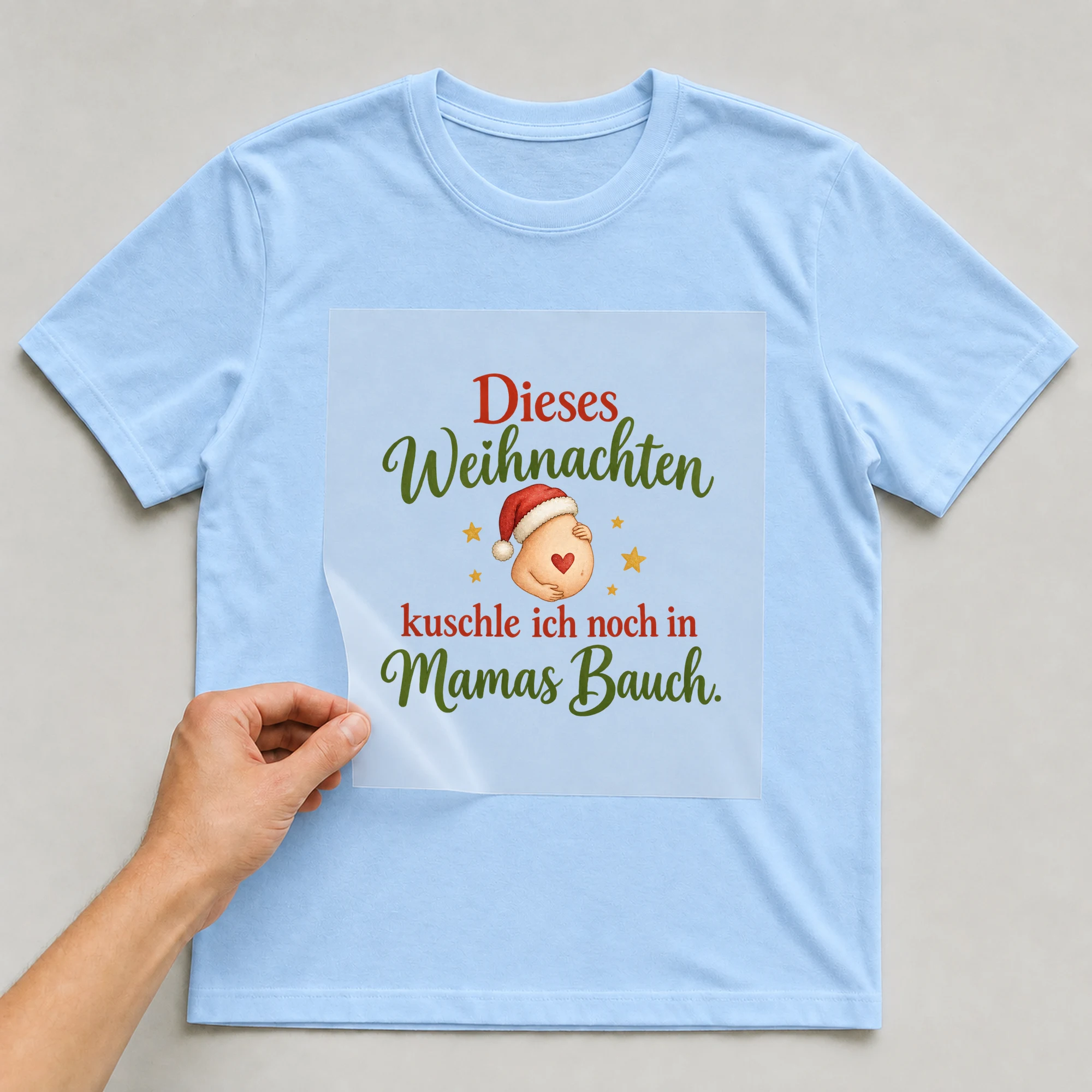 Bügelbild „Dieses Weihnachten kuschle ich noch in Mamas Bauch.“ auf babyblauem T-Shirt, Folie wird aufgelegt