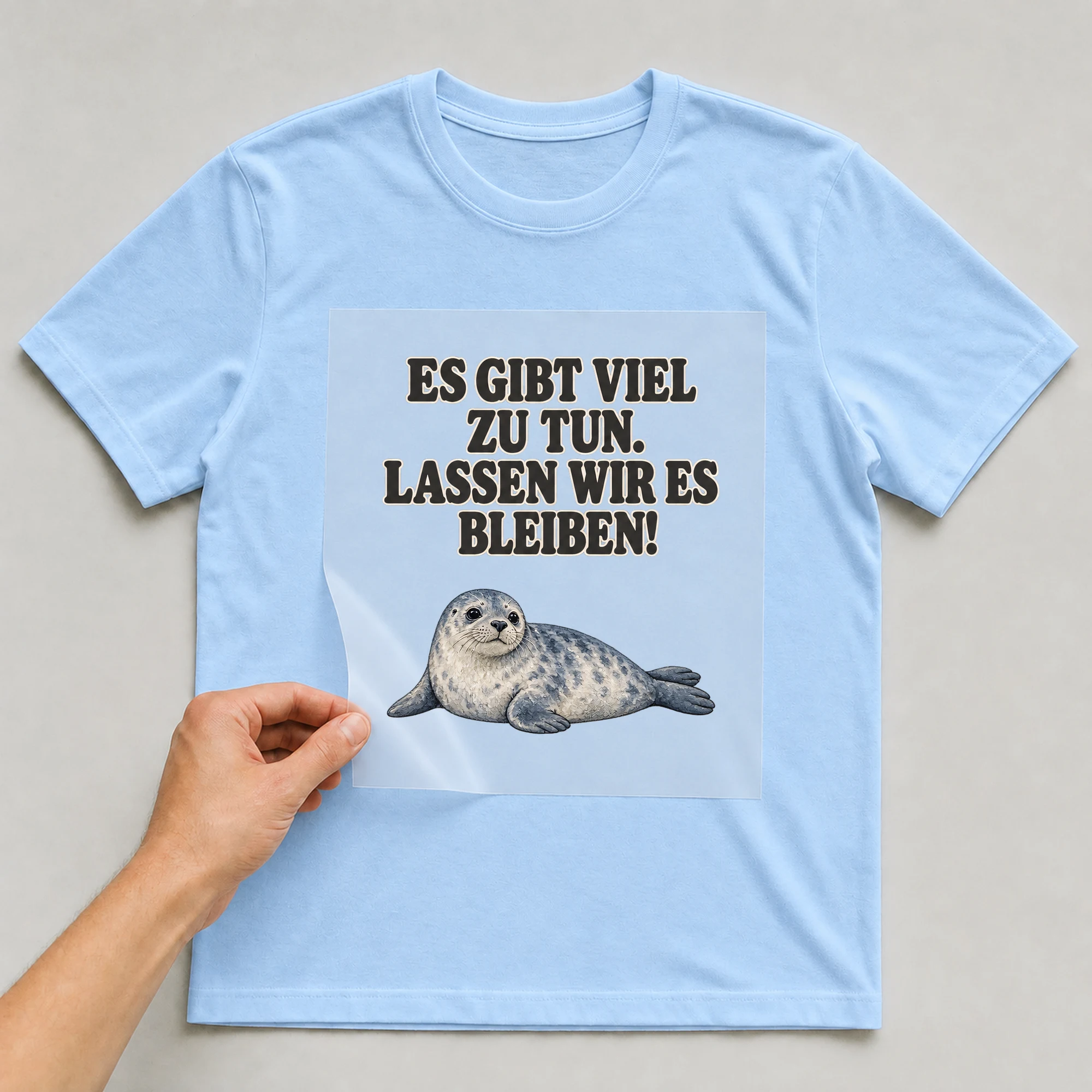 Bügelbild „ES GIBT VIEL ZU TUN. LASSEN WIR ES BLEIBEN!“ auf babyblauem T-Shirt, Folie wird aufgelegt