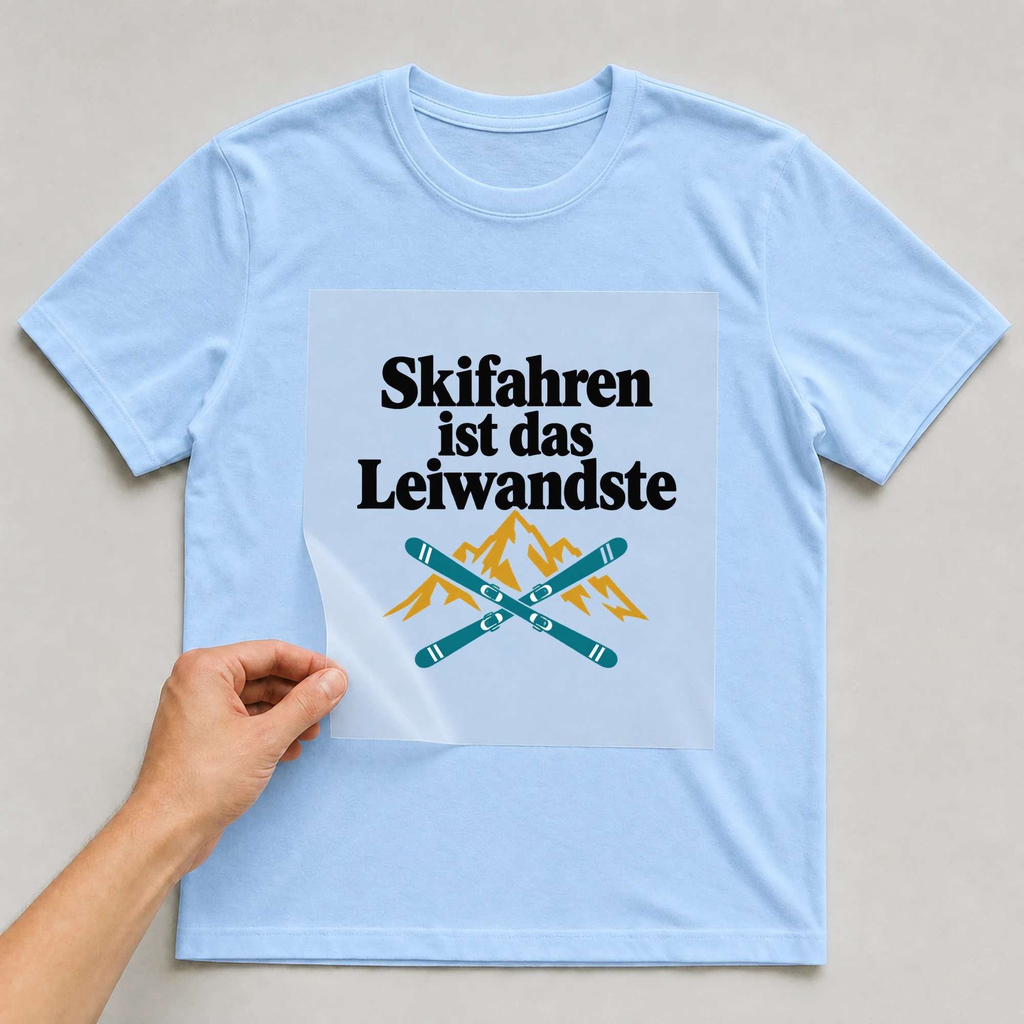Bügelbild „Skifahren ist das Leiwandste“ auf babyblauem T-Shirt, Folie wird aufgelegt
