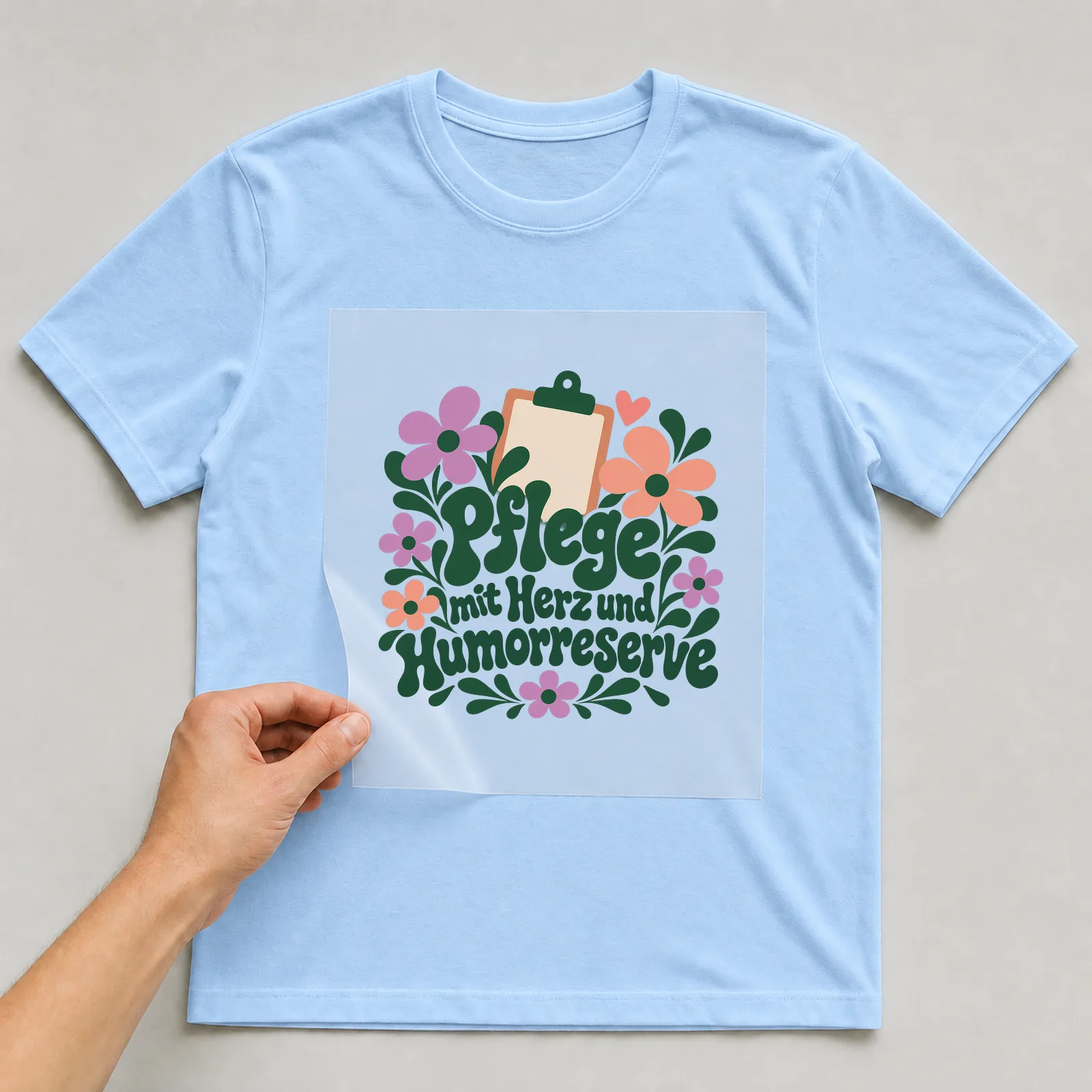 Bügelbild „Pflege mit Herz und Humorreserve“ auf hellblauem T-Shirt, Folie wird aufgelegt