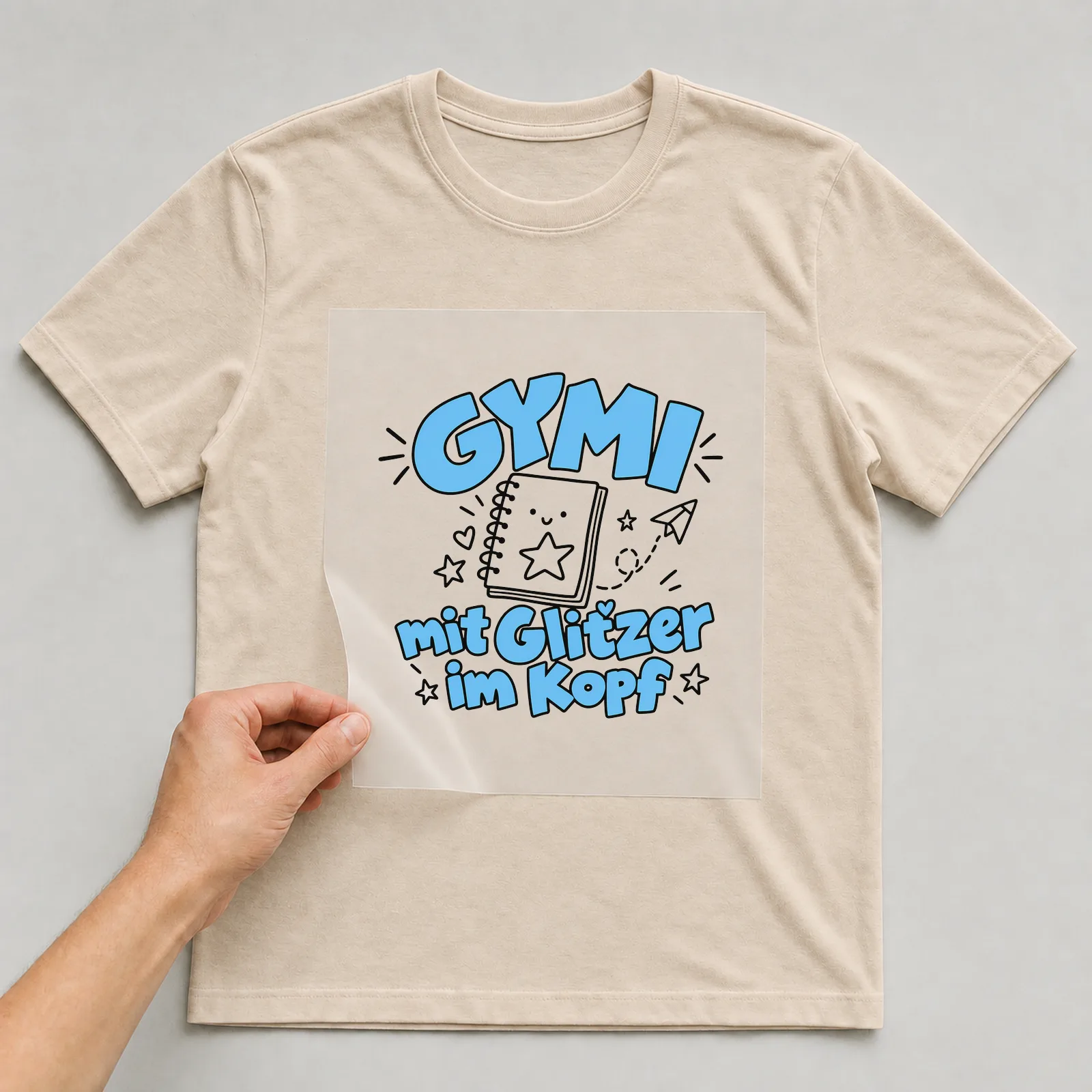 Bügelbild „Gymi mit Glitzer im Kopf“ auf beigem T-Shirt, Folie wird aufgelegt