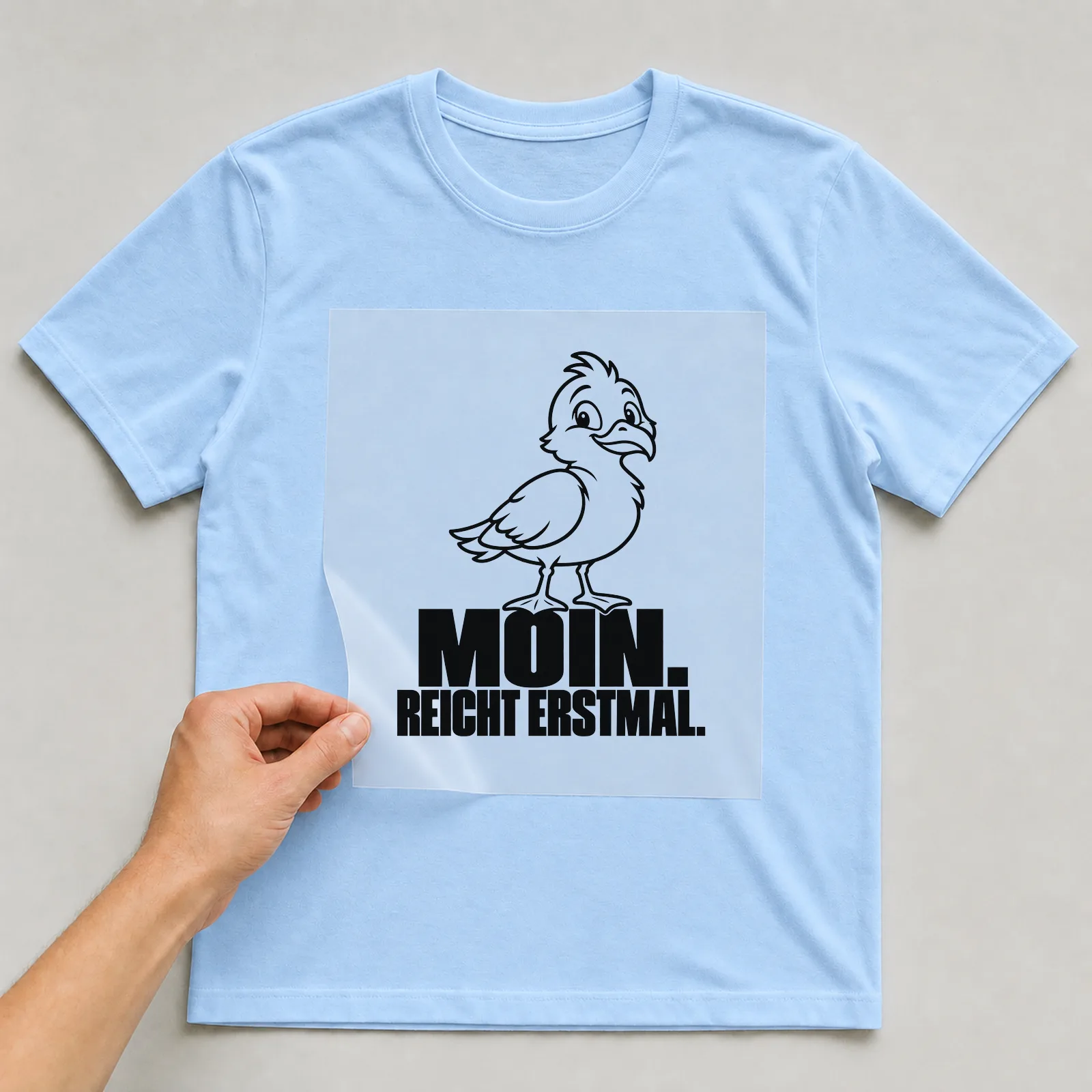 Bügelbild „Moin. Reicht Erstmal.“ auf hellblauem T-Shirt, Folie wird aufgelegt