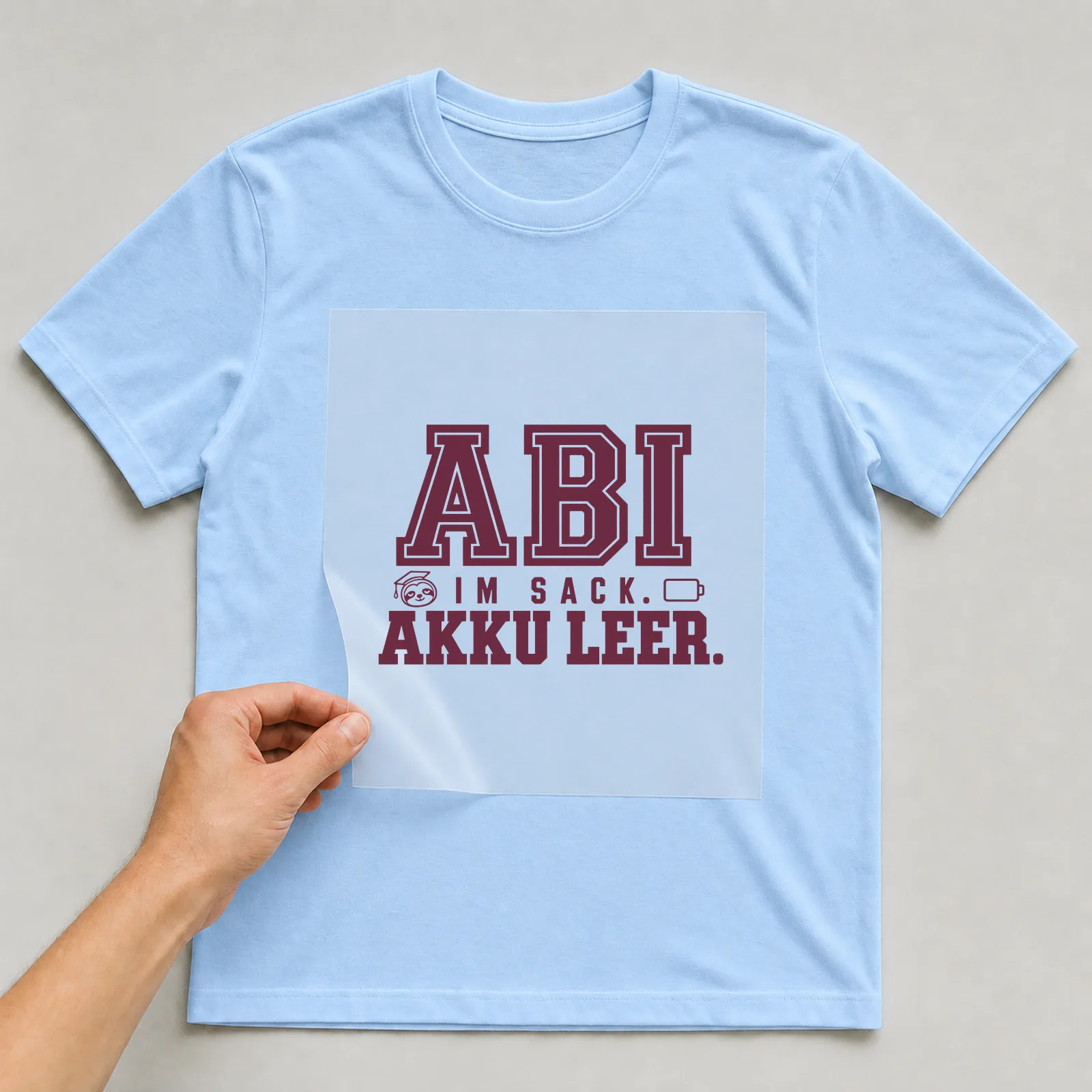 Bügelbild „Abi im Sack. Akku leer.“ (Schrift Konturen) auf hellblauem T-Shirt, Folie wird aufgelegt