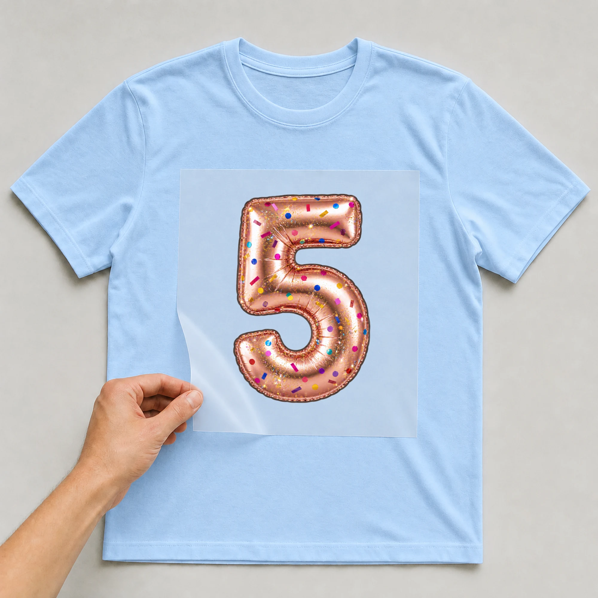 Bügelbild „Zahl 5 im Konfetti-Metallic-Look“ auf babyblauem T-Shirt, Folie wird aufgelegt