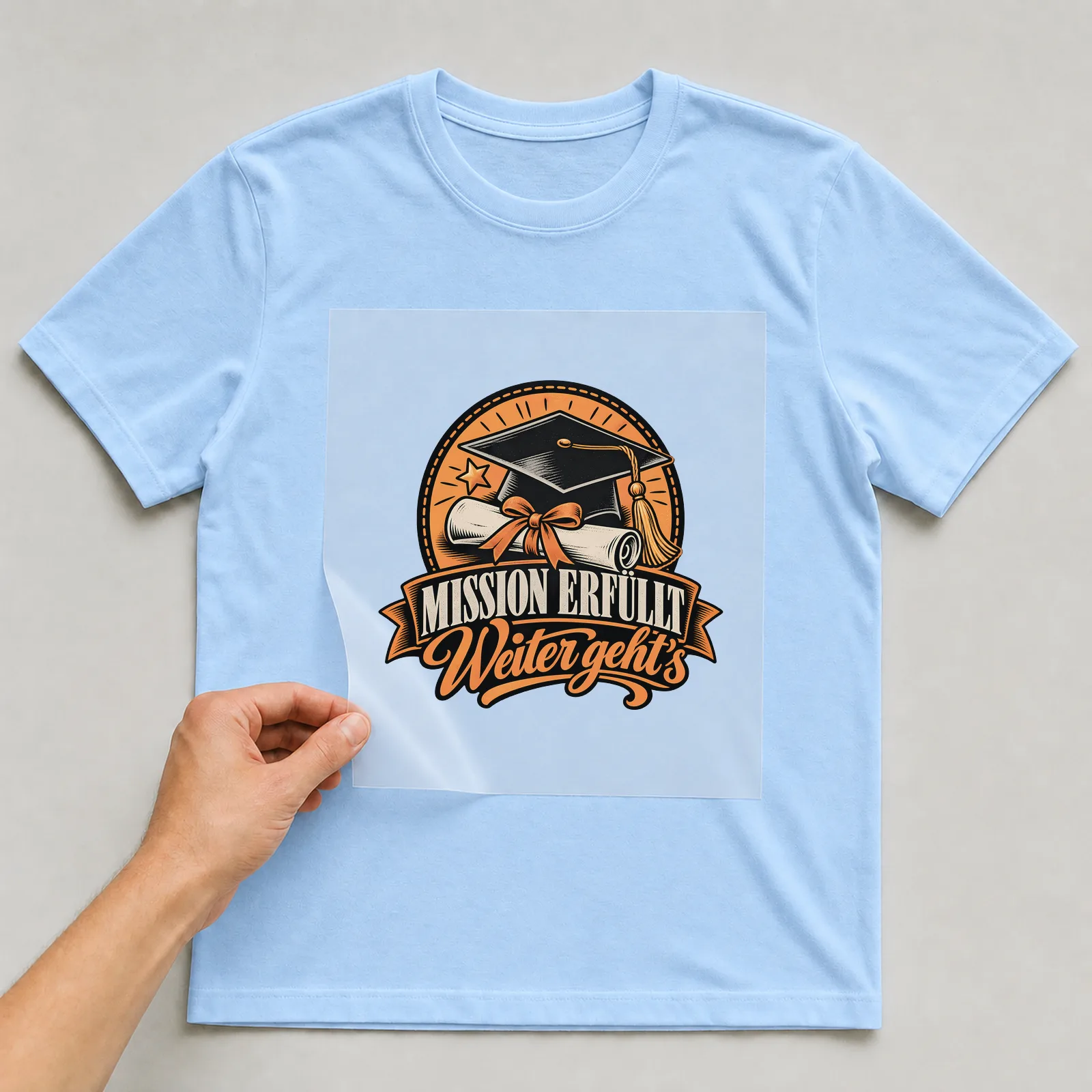 Bügelbild „Mission erfüllt Weiter geht’s“ auf hellblauem T-Shirt, Folie wird aufgelegt