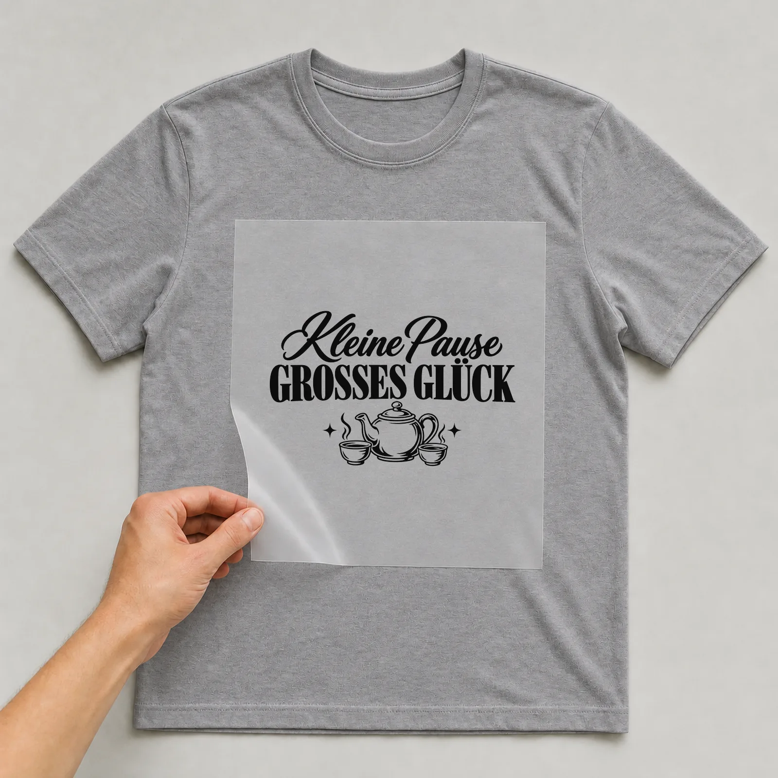 Bügelbild „Kleine Pause · GROSSES GLÜCK“ auf grauem T-Shirt, Folie wird aufgelegt