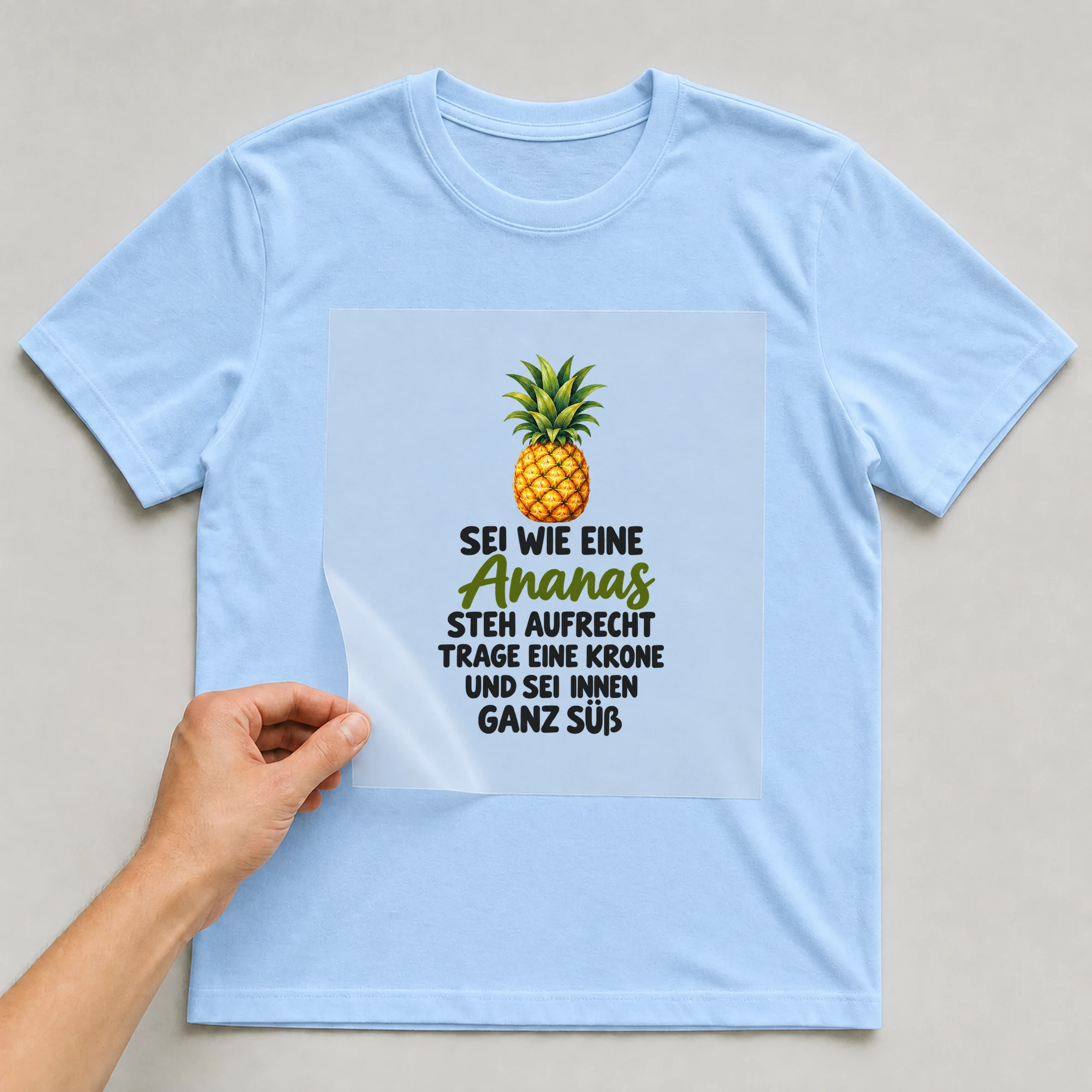 Bügelbild „Sei wie eine Ananas steh aufrecht trage eine Krone und SEI innen ganz süß“ auf babyblauem T-Shirt, Folie wird aufgelegt