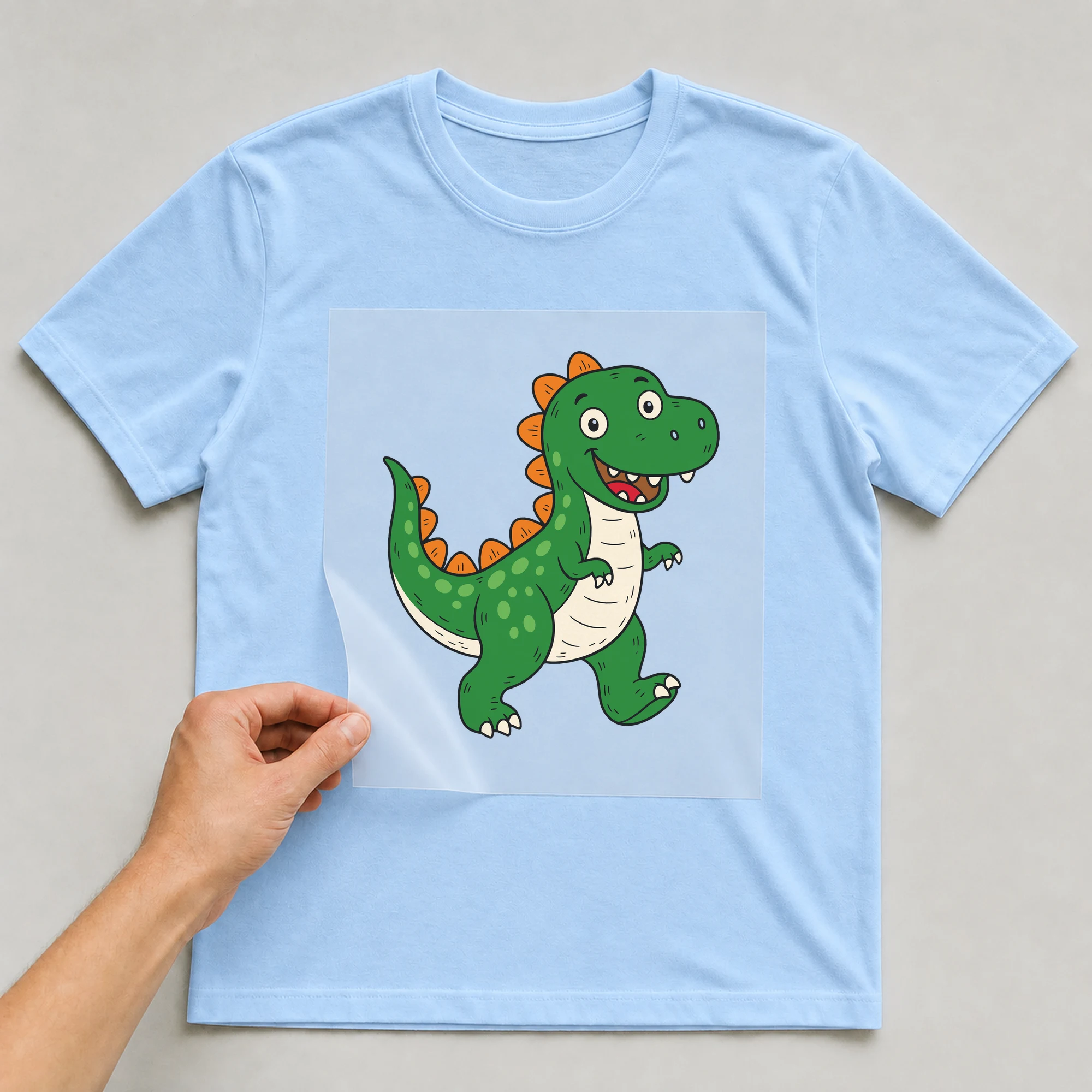 Bügelbild „Dino T-Rex“ auf babyblauem T-Shirt, Folie wird aufgelegt