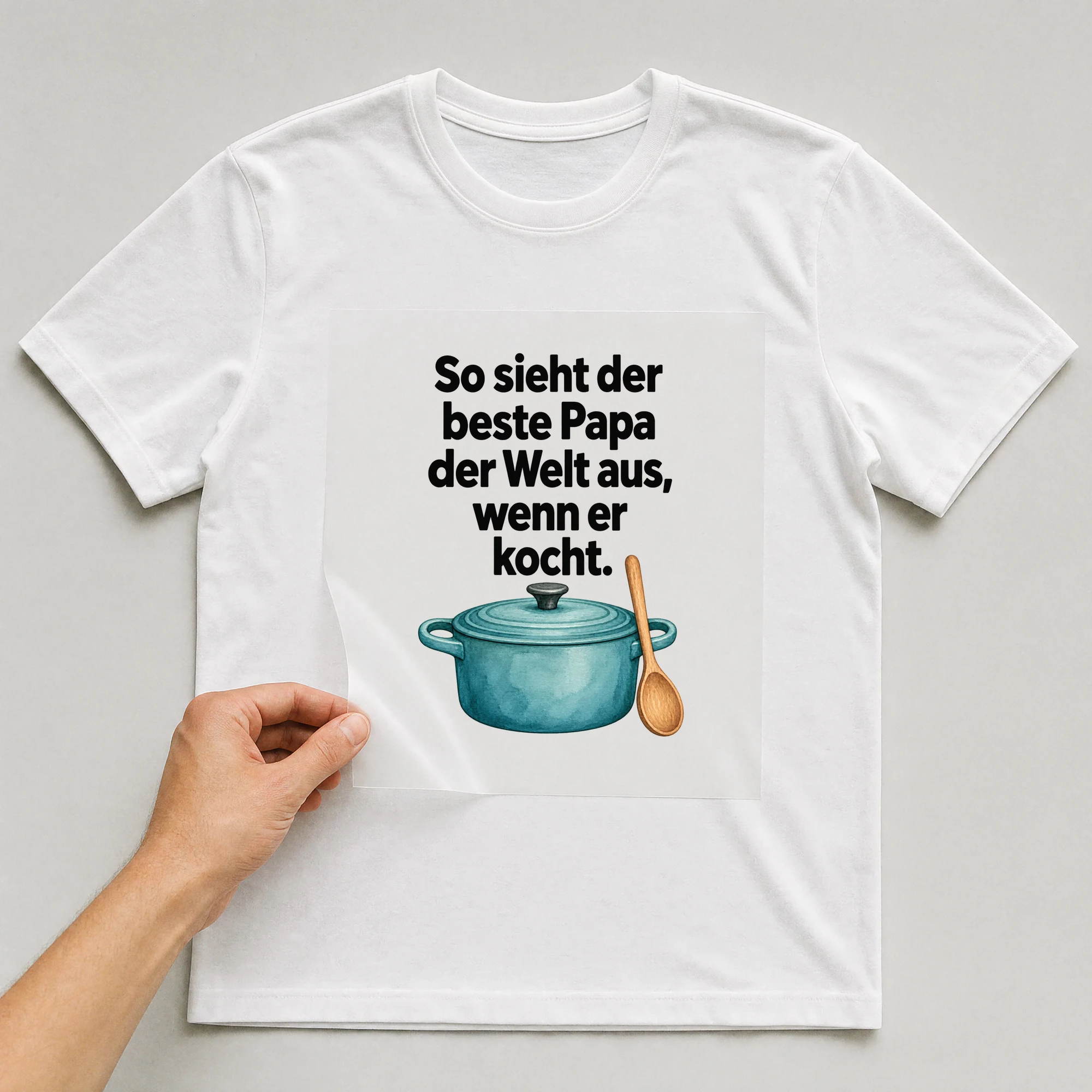 Bügelbild „So sieht der beste Papa der Welt aus, wenn er kocht.“ auf weißem T-Shirt, Folie wird aufgelegt