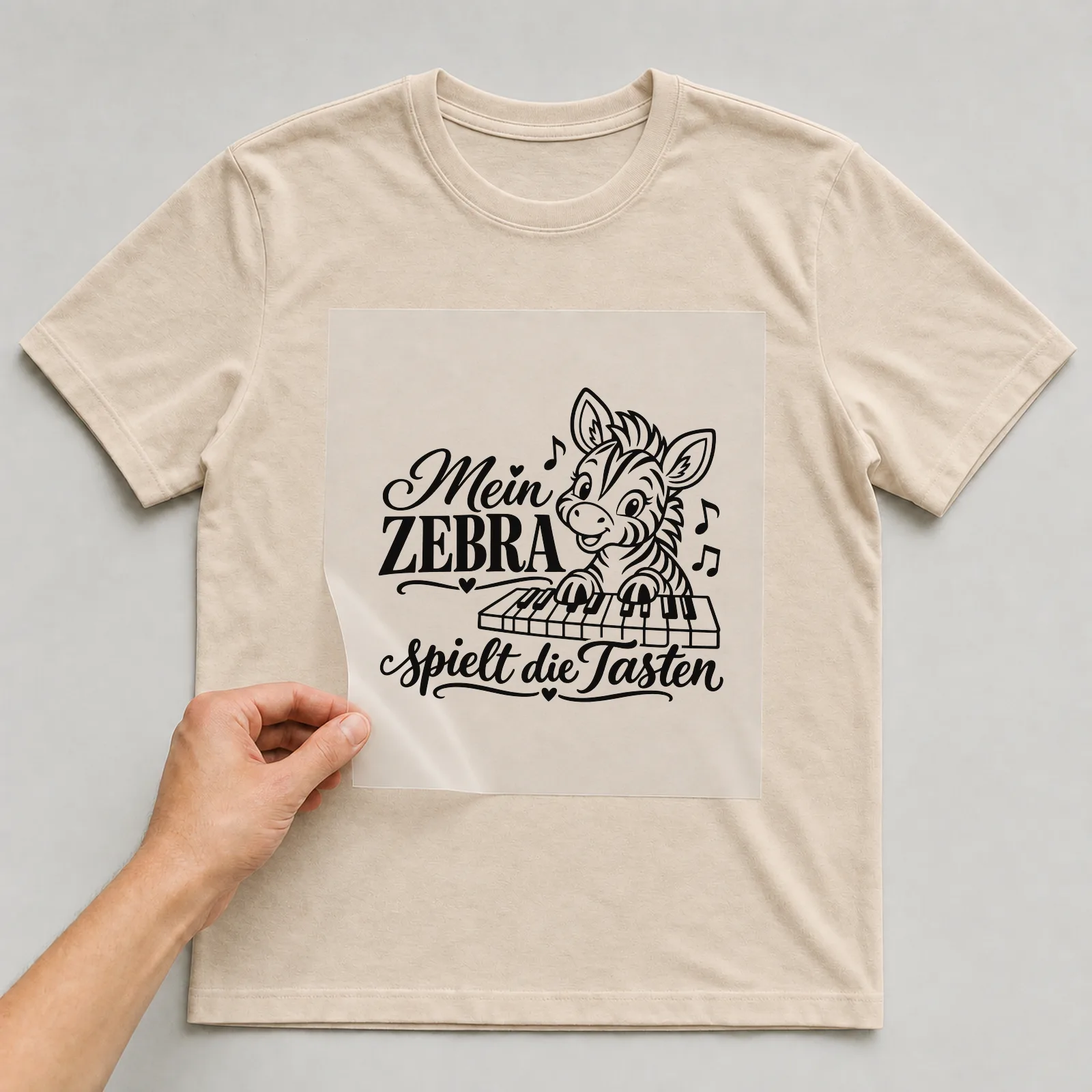 Bügelbild „Mein Zebra spielt die Tasten“ auf beigem T-Shirt, Folie wird aufgelegt