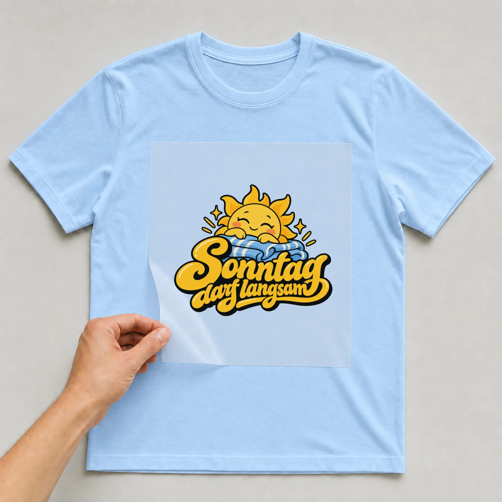 Bügelbild „Sonntag darf langsam“ auf hellblauem T-Shirt, Folie wird aufgelegt