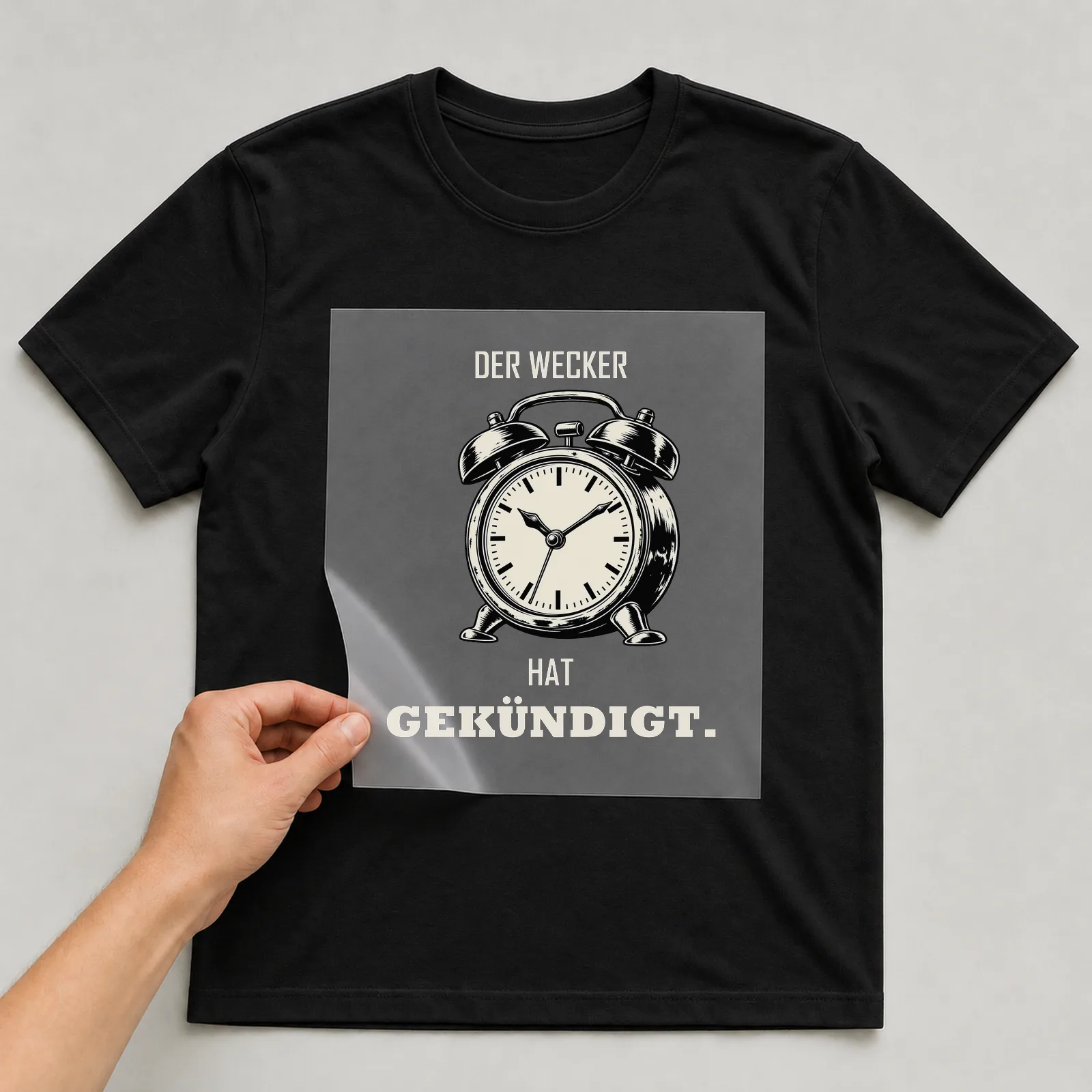 Bügelbild „Der Wecker hat gekündigt“ auf schwarzem T-Shirt, Folie wird aufgelegt