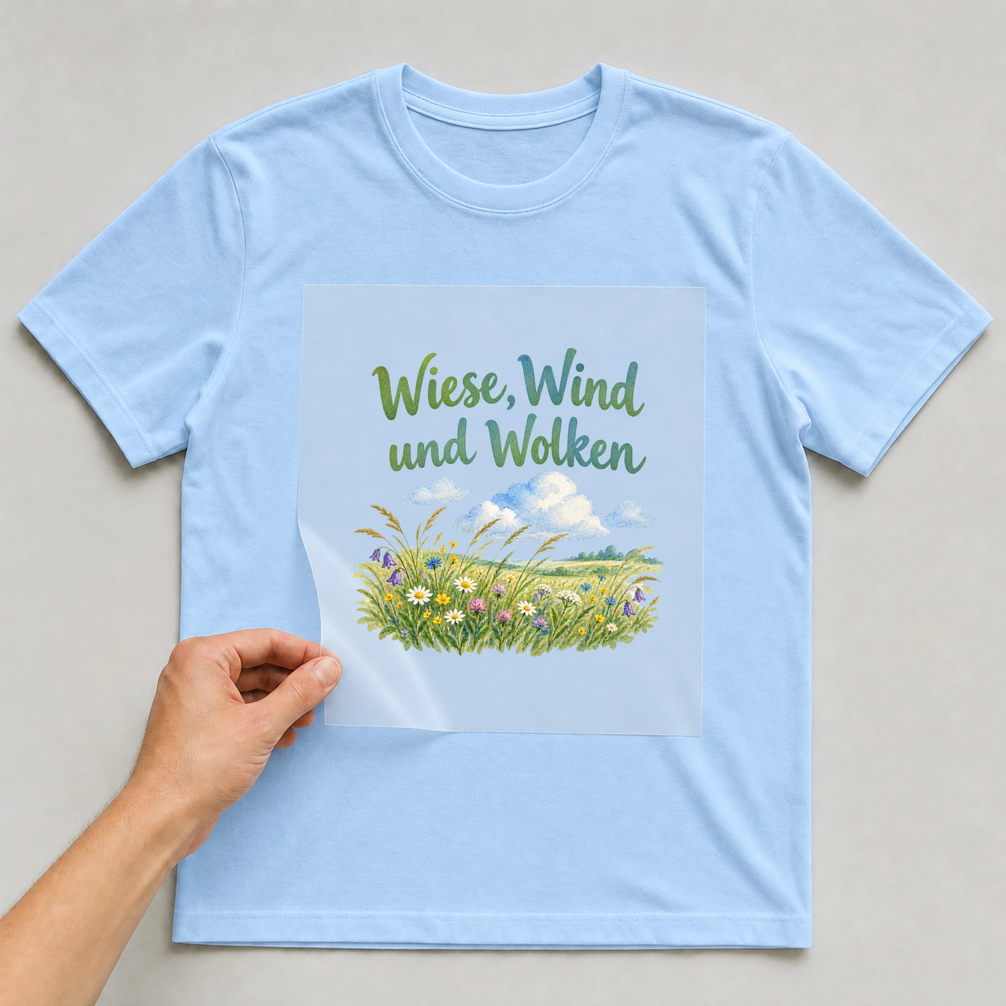 Bügelbild „Wiese, Wind und Wolken“ auf babyblauem T-Shirt, Folie wird aufgelegt