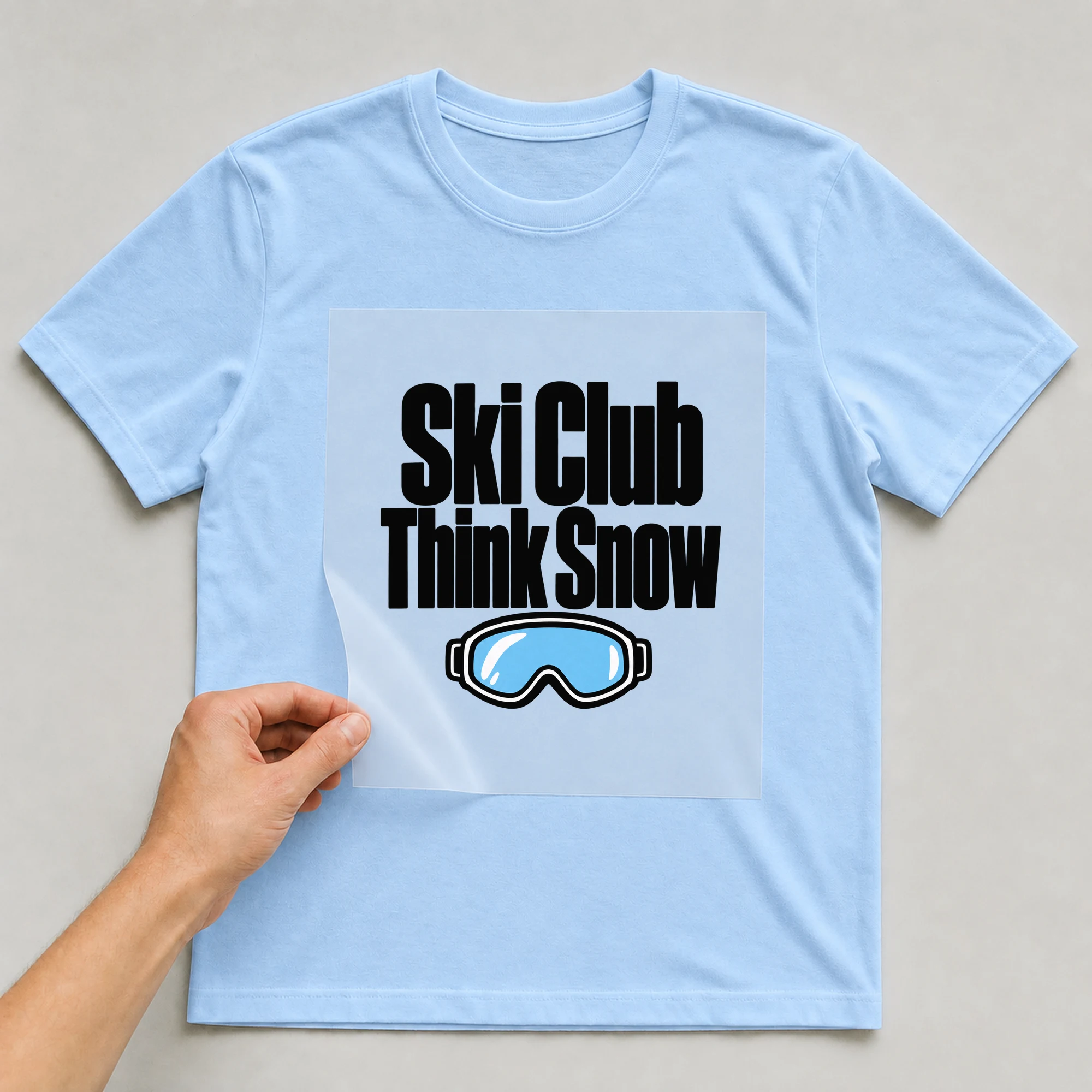 Bügelbild „Ski Club Think Snow“ auf babyblauem T-Shirt, Folie wird aufgelegt