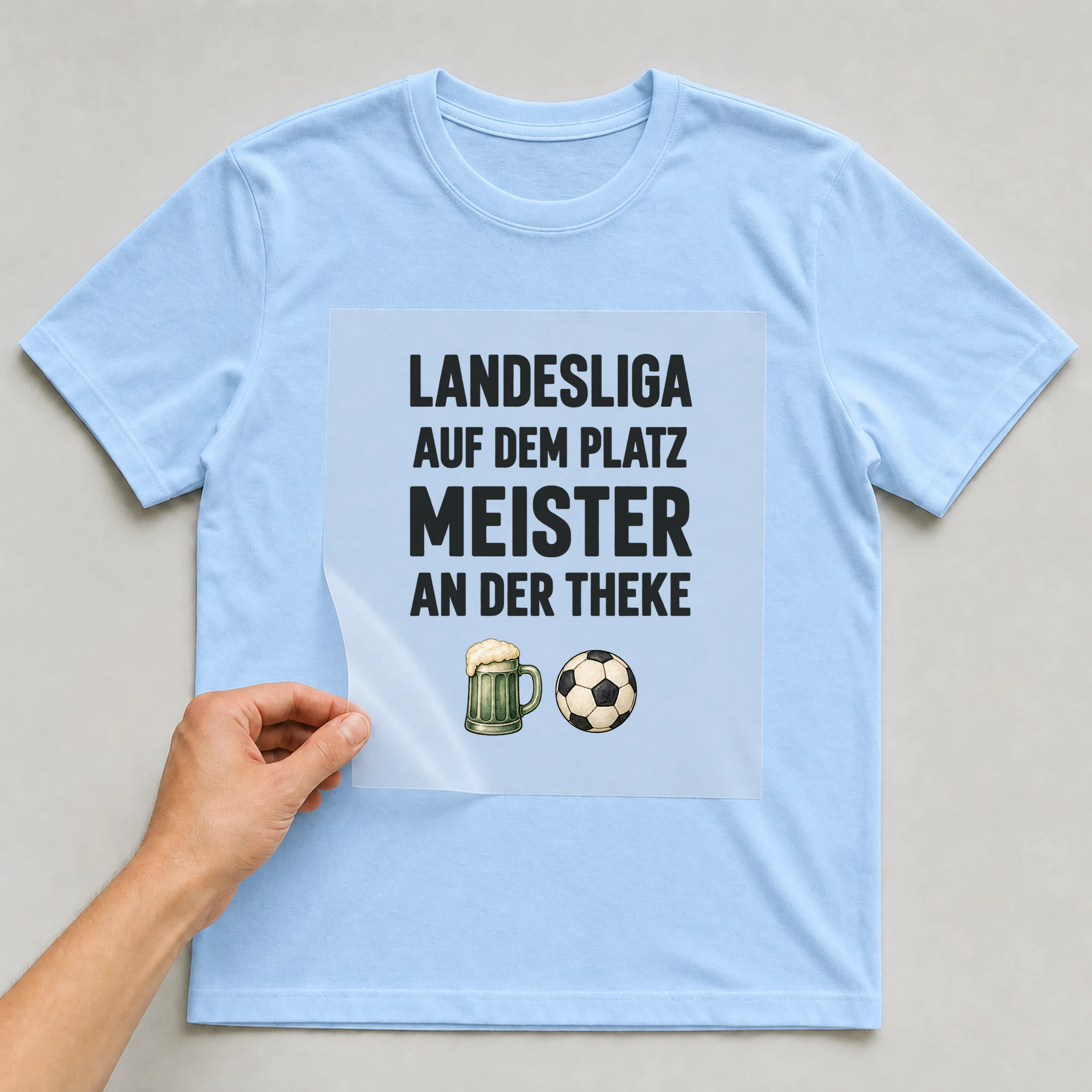 Bügelbild „LANDESLIGA AUF DEM PLATZ MEISTER AN DER THEKE“ auf babyblauem T-Shirt, Folie wird aufgelegt