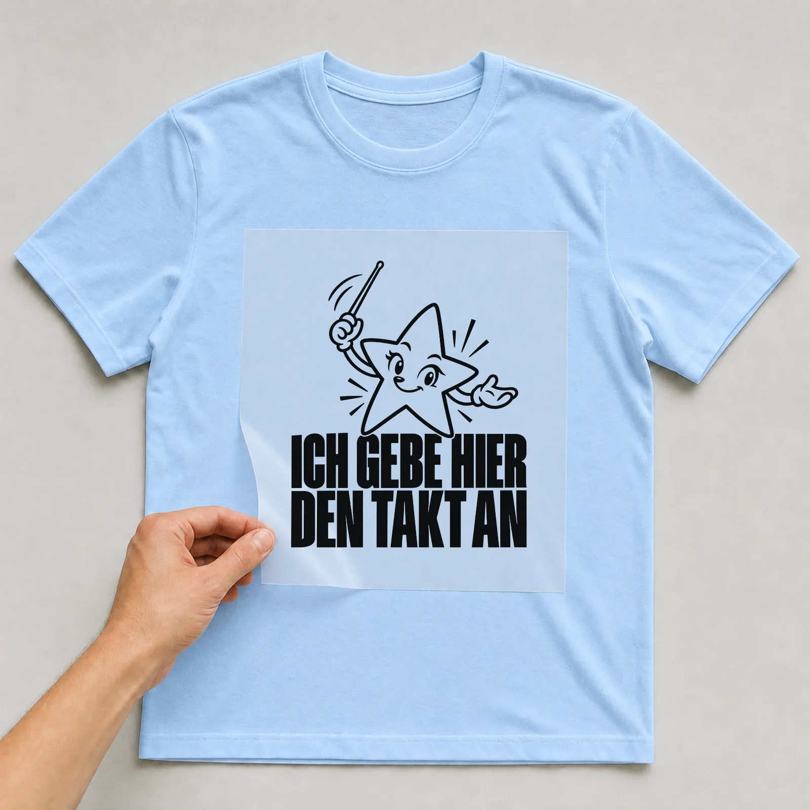 Bügelbild „Ich gebe hier den Takt an“ auf hellblauem T-Shirt, Folie wird aufgelegt