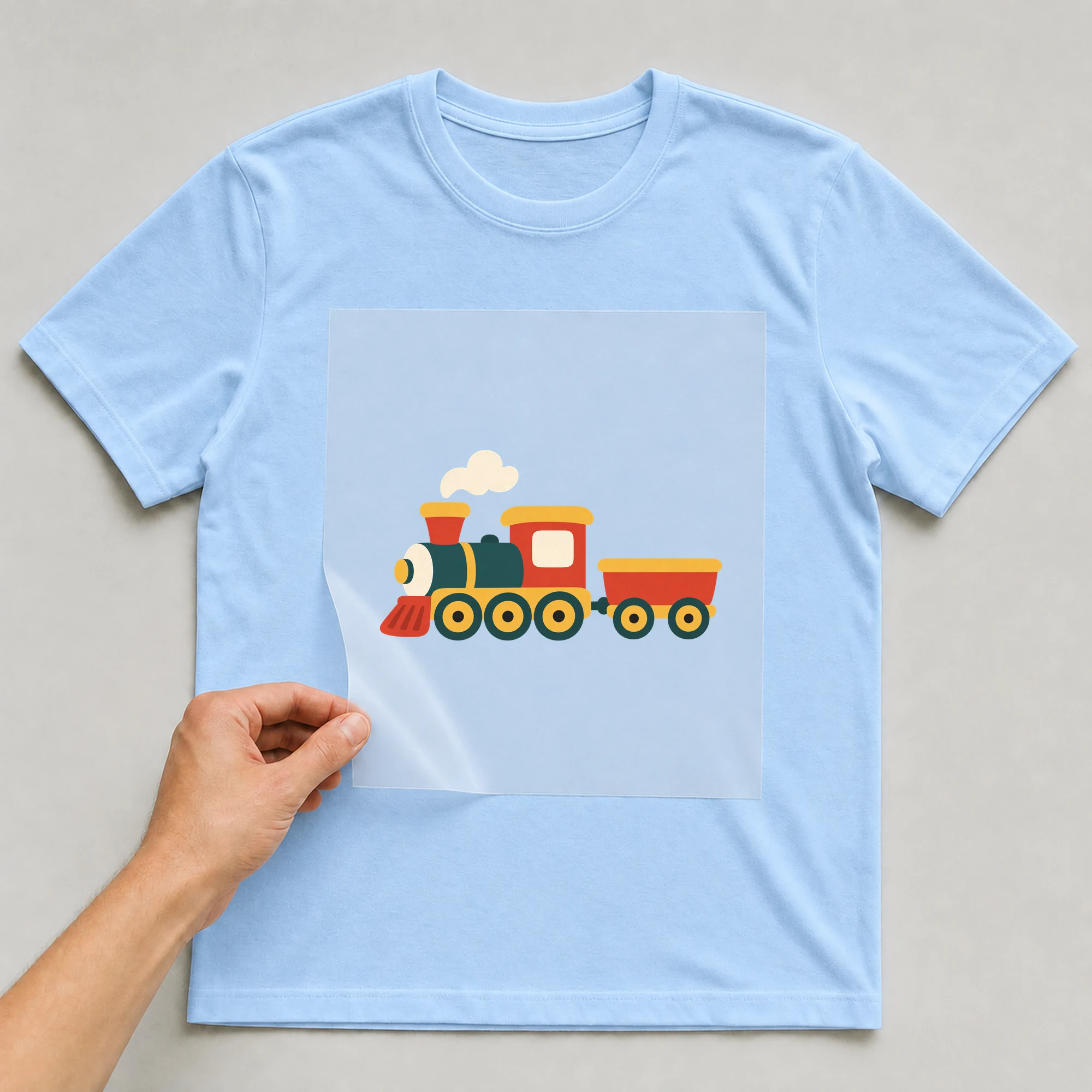 Bügelbild „Baby Comic - Lokomotive“ auf babyblauem T-Shirt, Folie wird aufgelegt