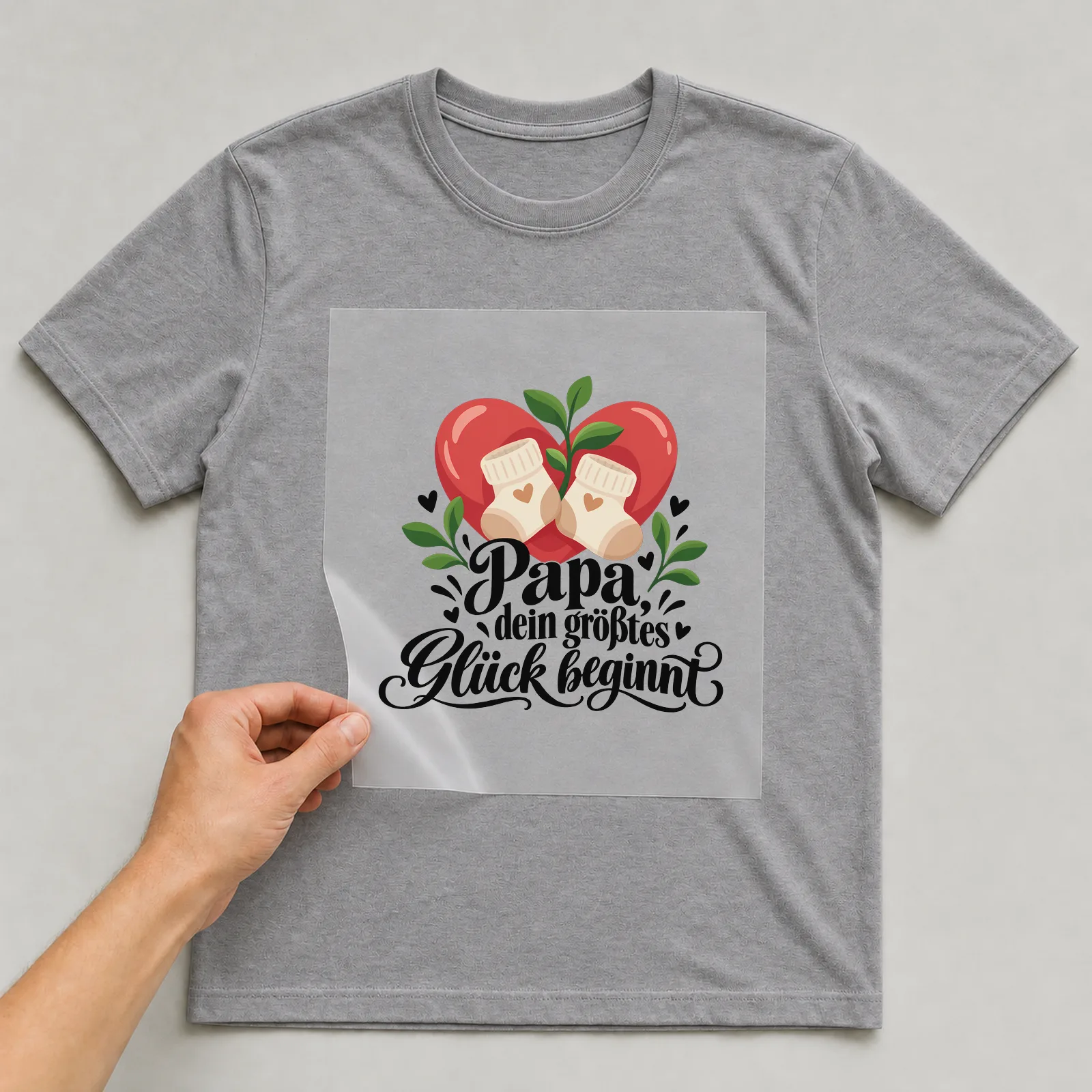 Bügelbild „Papa, dein größtes Glück beginnt“ auf grauem T-Shirt, Folie wird aufgelegt