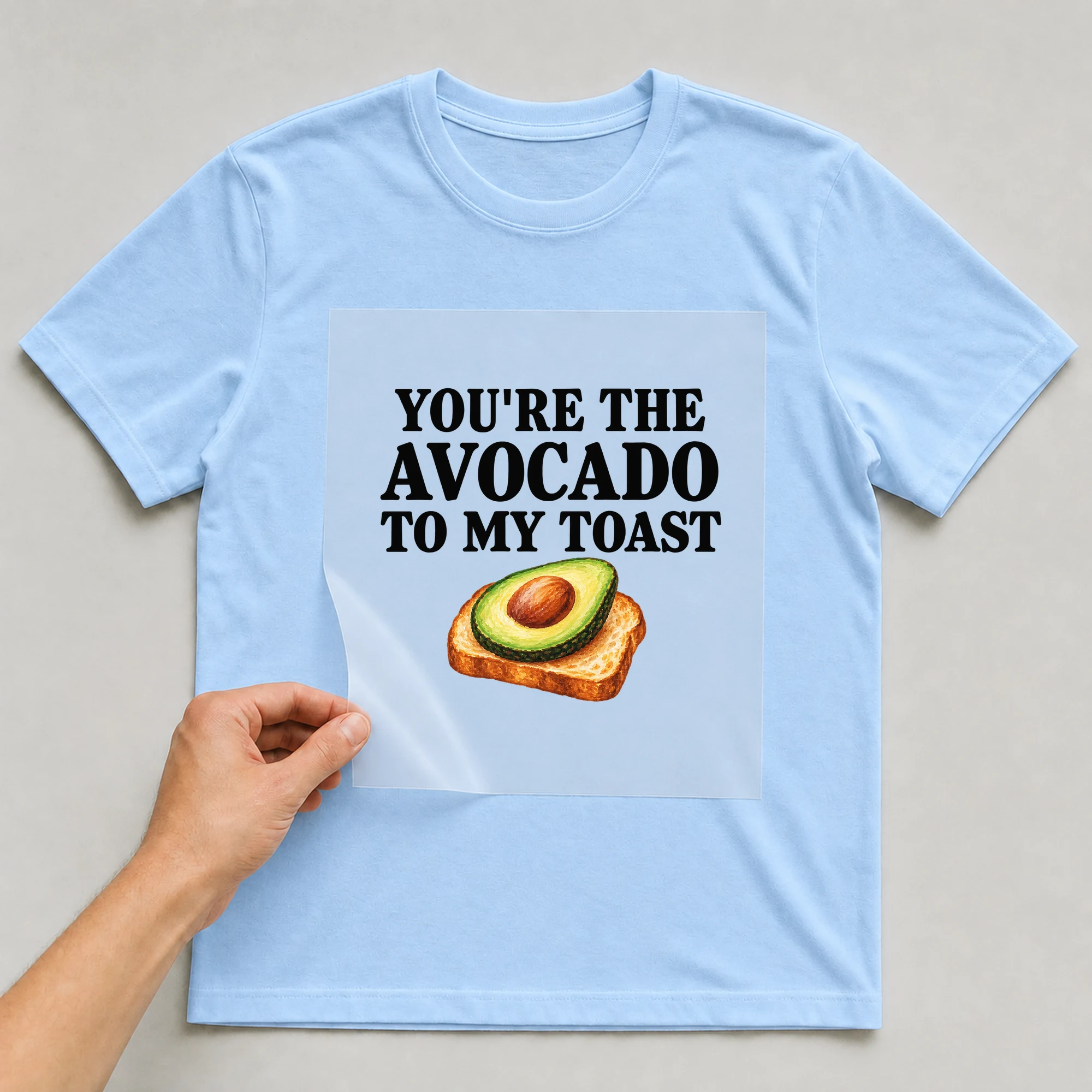 Bügelbild „You're The Avocado To My Toast“ auf babyblauem T-Shirt, Folie wird aufgelegt