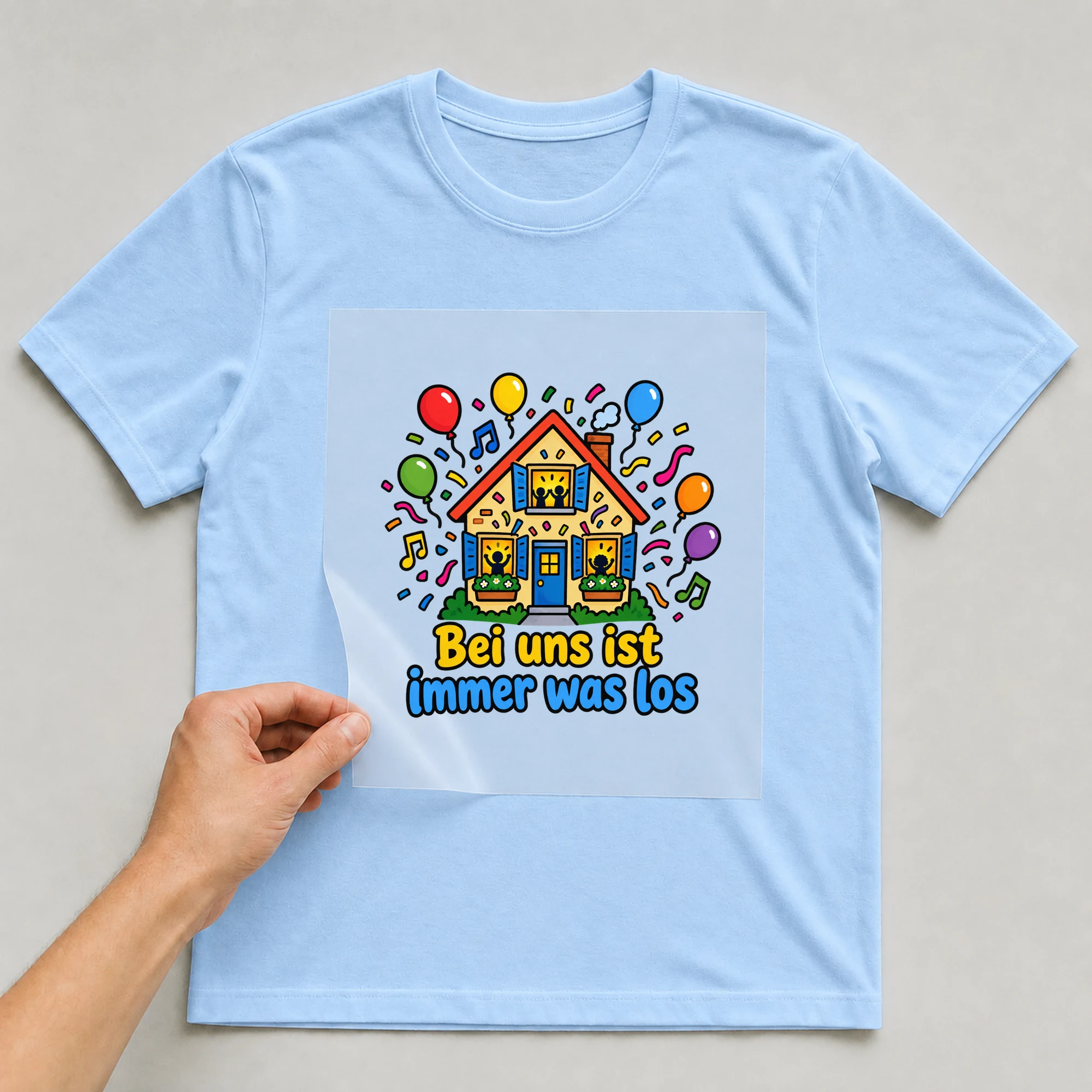 Bügelbild „Bei uns ist immer was los“ auf babyblauem T-Shirt, Folie wird aufgelegt
