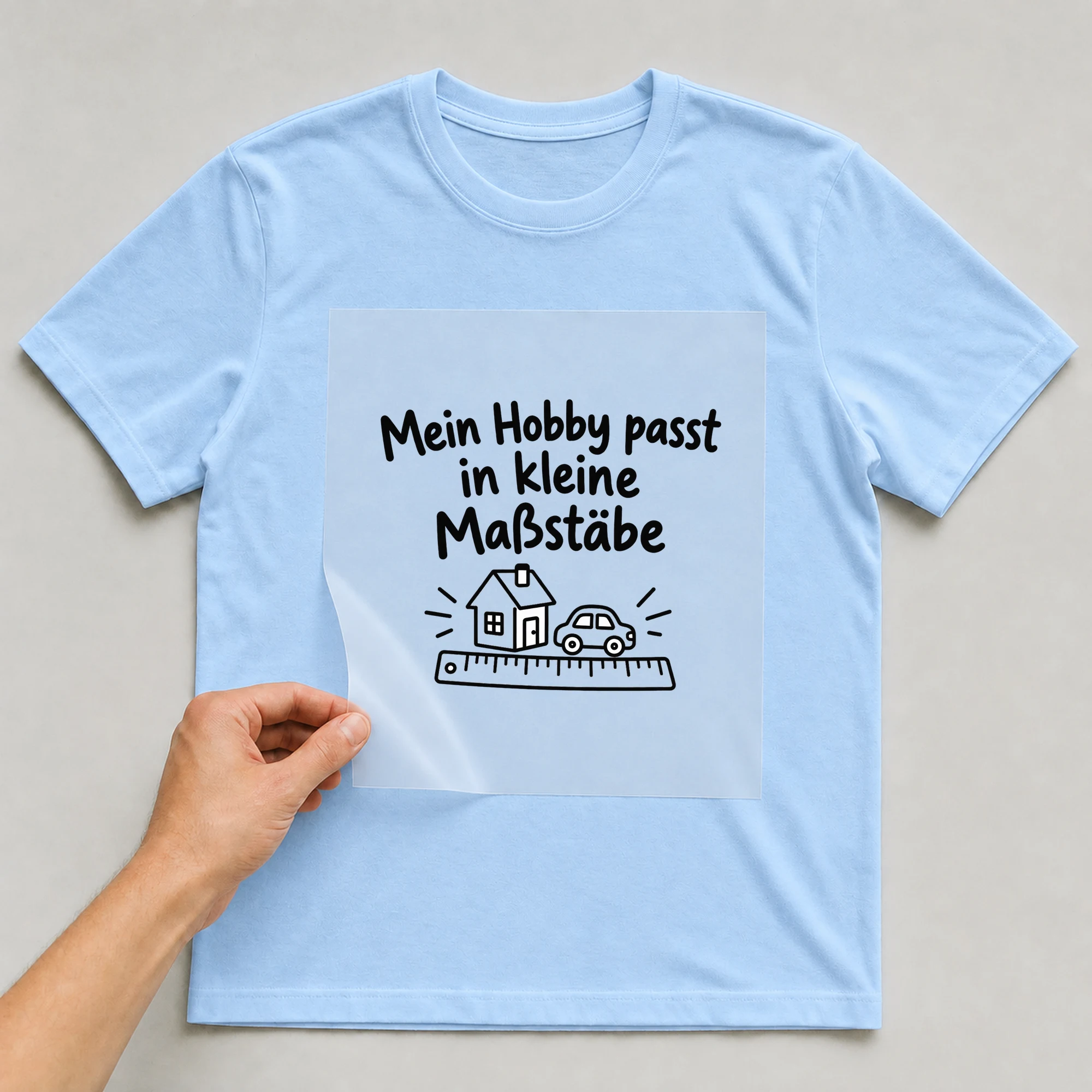 Bügelbild „Mein Hobby passt in kleine Maßstäbe“ auf babyblauem T-Shirt, Folie wird aufgelegt