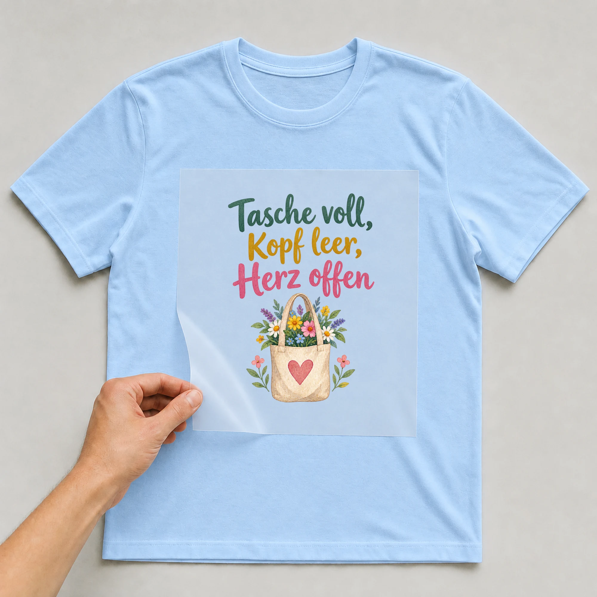 Bügelbild „Tasche voll, Kopf leer, Herz offen“ auf babyblauem T-Shirt, Folie wird aufgelegt