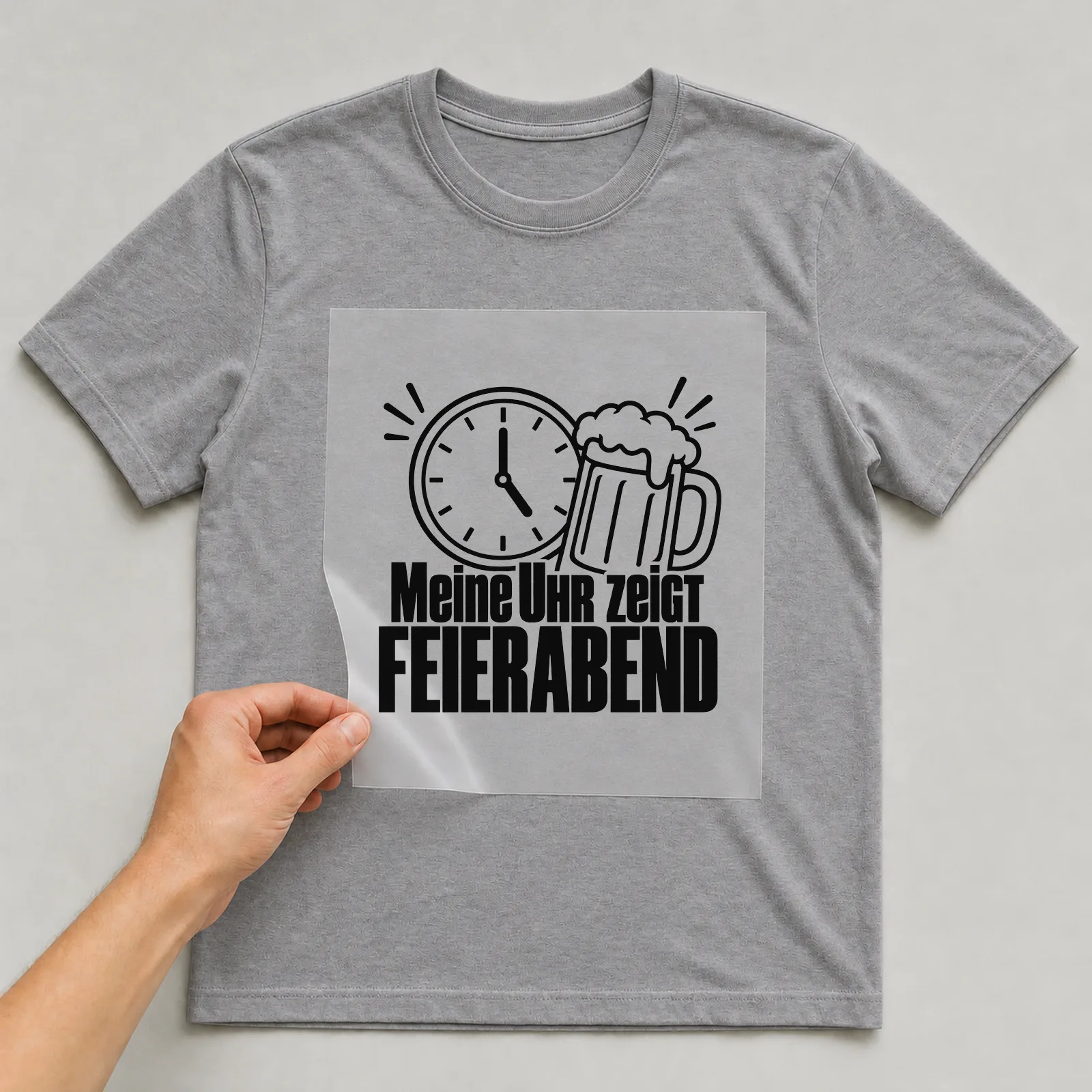 Bügelbild „Meine Uhr zeigt FEIERABEND“ auf grauem T-Shirt, Folie wird aufgelegt