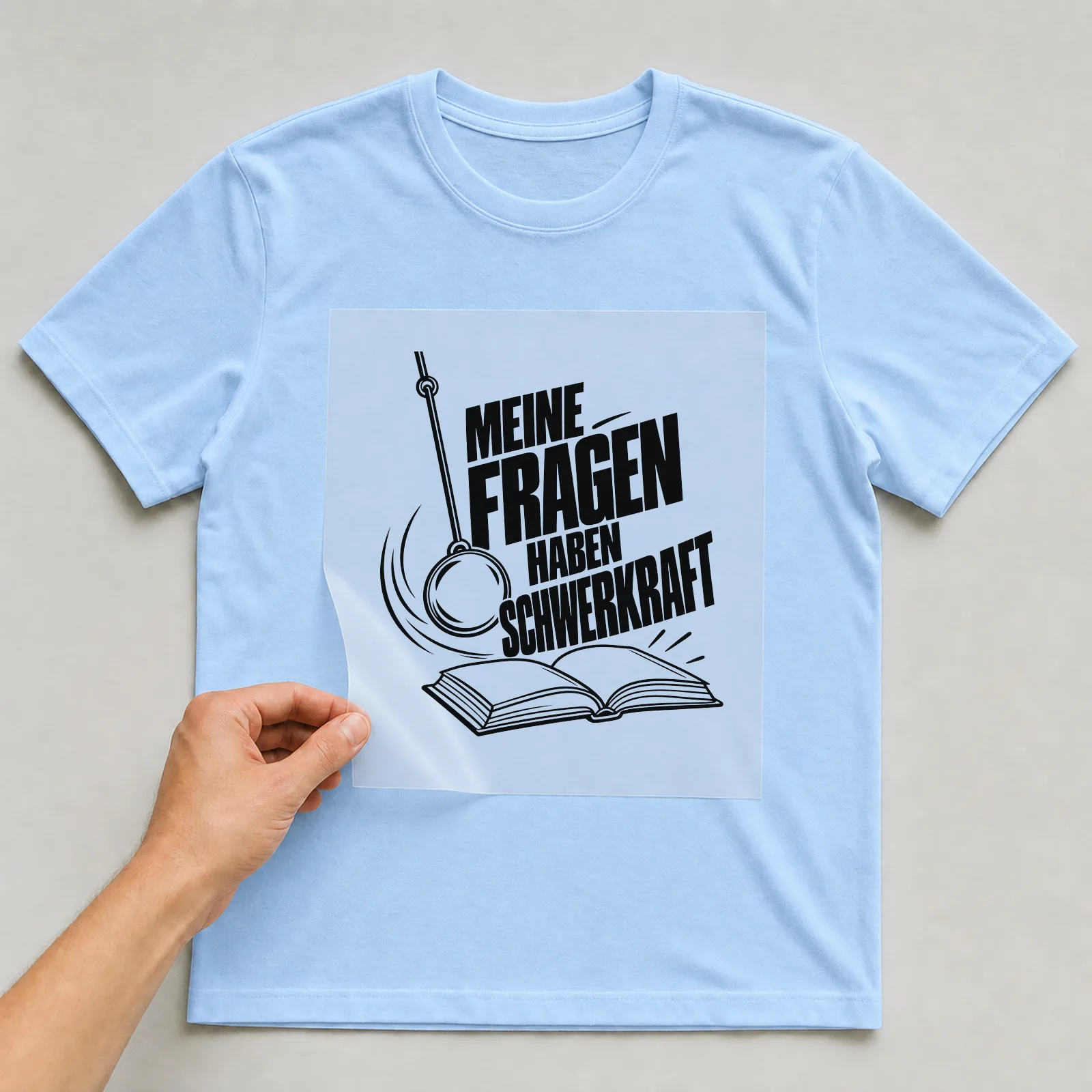 Bügelbild „Meine Fragen haben Schwerkraft“ auf hellblauem T-Shirt, Folie wird aufgelegt
