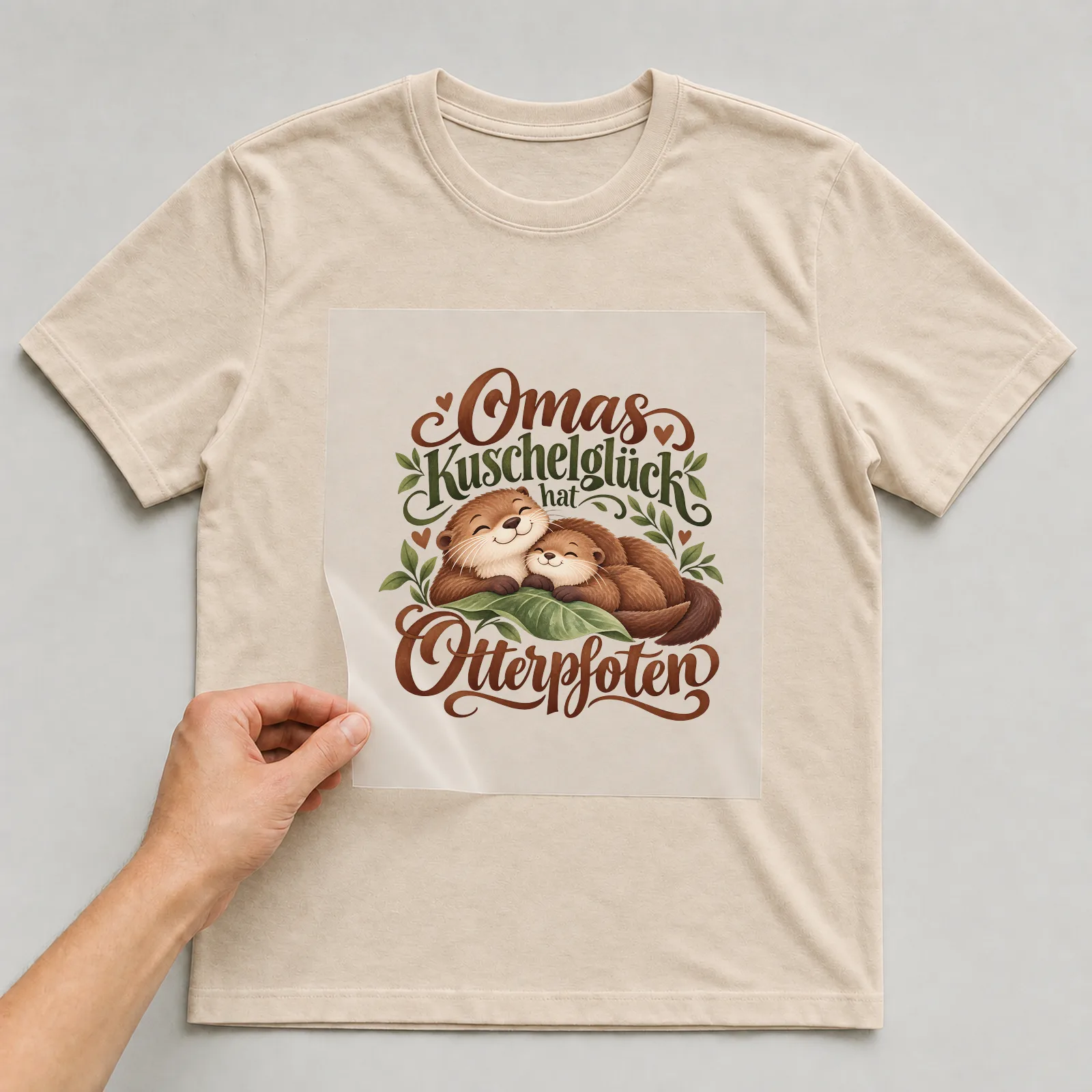 Bügelbild „Omas Kuschelglück hat Otterpfoten“ auf beigem T-Shirt, Folie wird aufgelegt