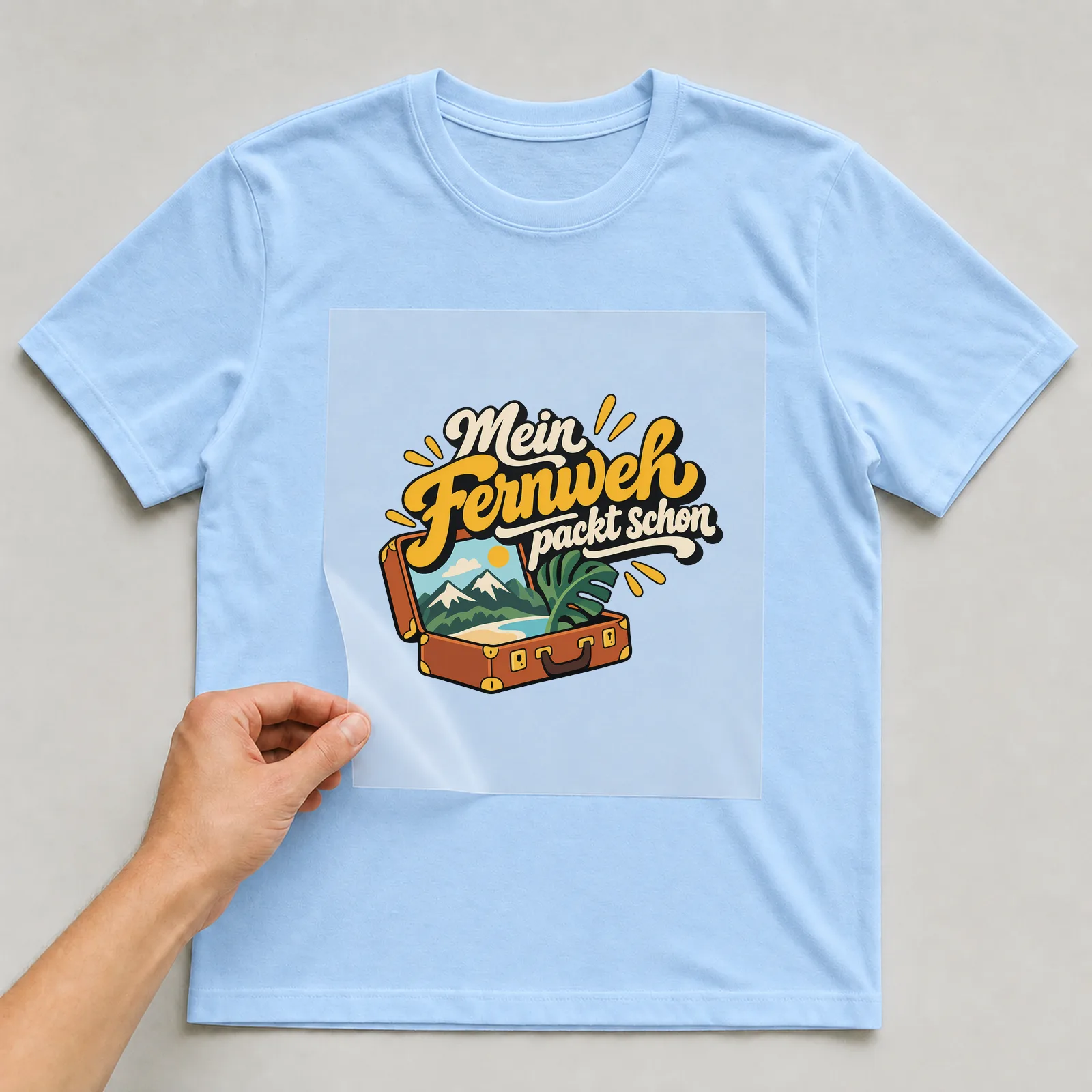 Bügelbild „Mein Fernweh packt schon“ auf hellblauem T-Shirt, Folie wird aufgelegt