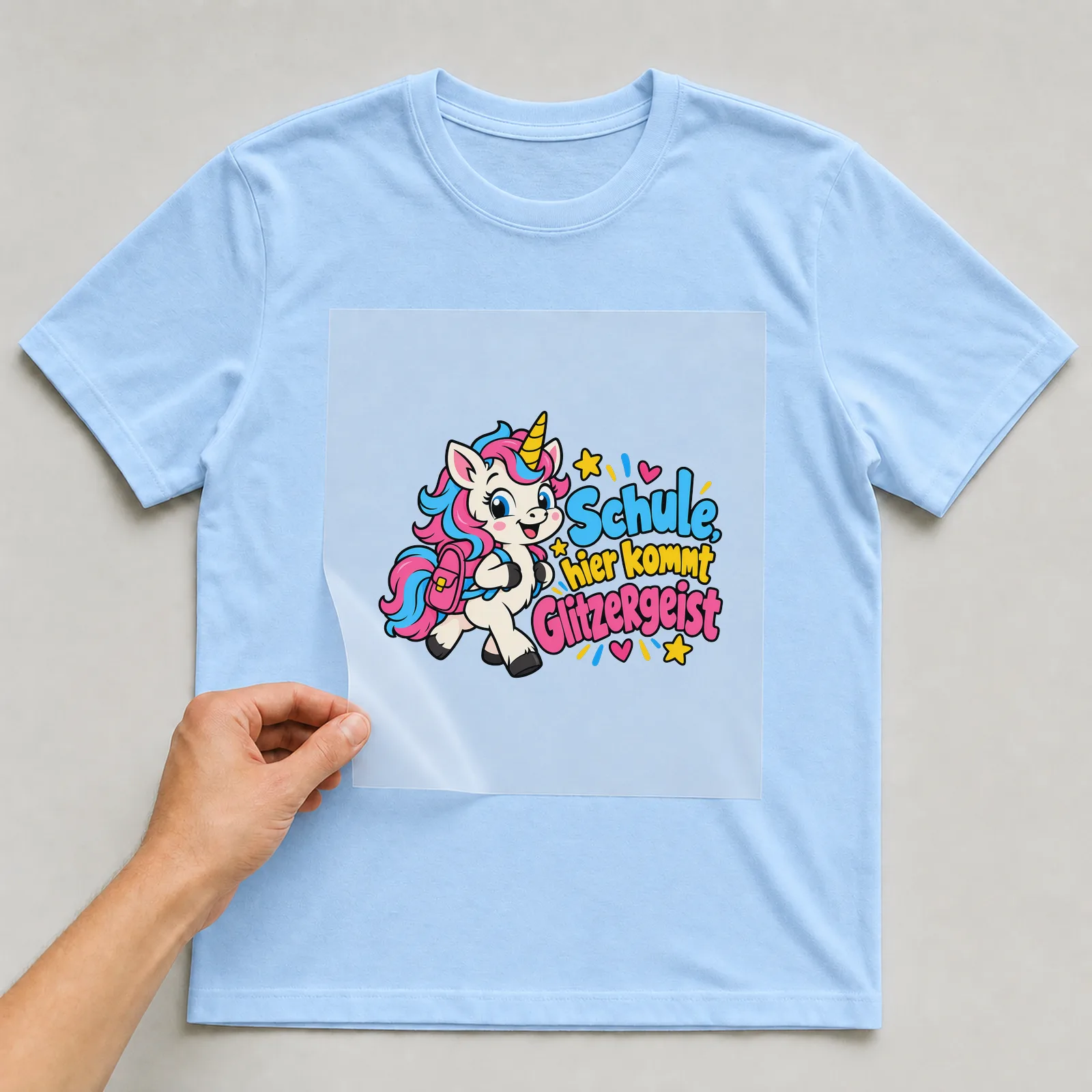 Bügelbild „Schule, hier kommt Glitzergeist“ auf hellblauem T-Shirt, Folie wird aufgelegt