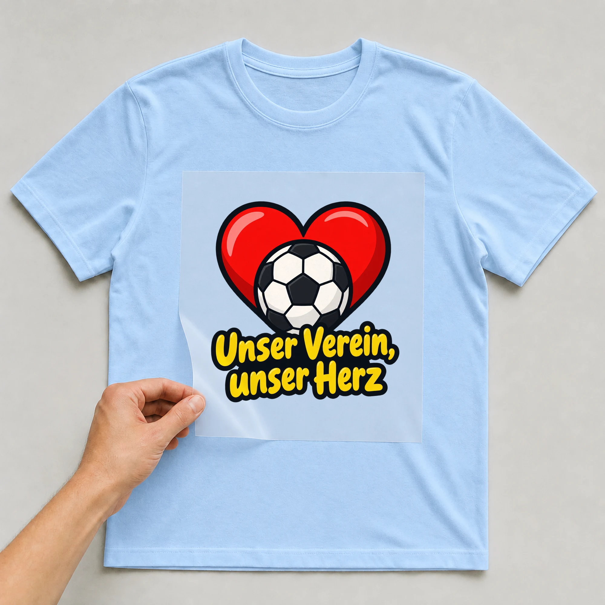 Bügelbild „Unser Verein, unser Herz“ auf babyblauem T-Shirt, Folie wird aufgelegt