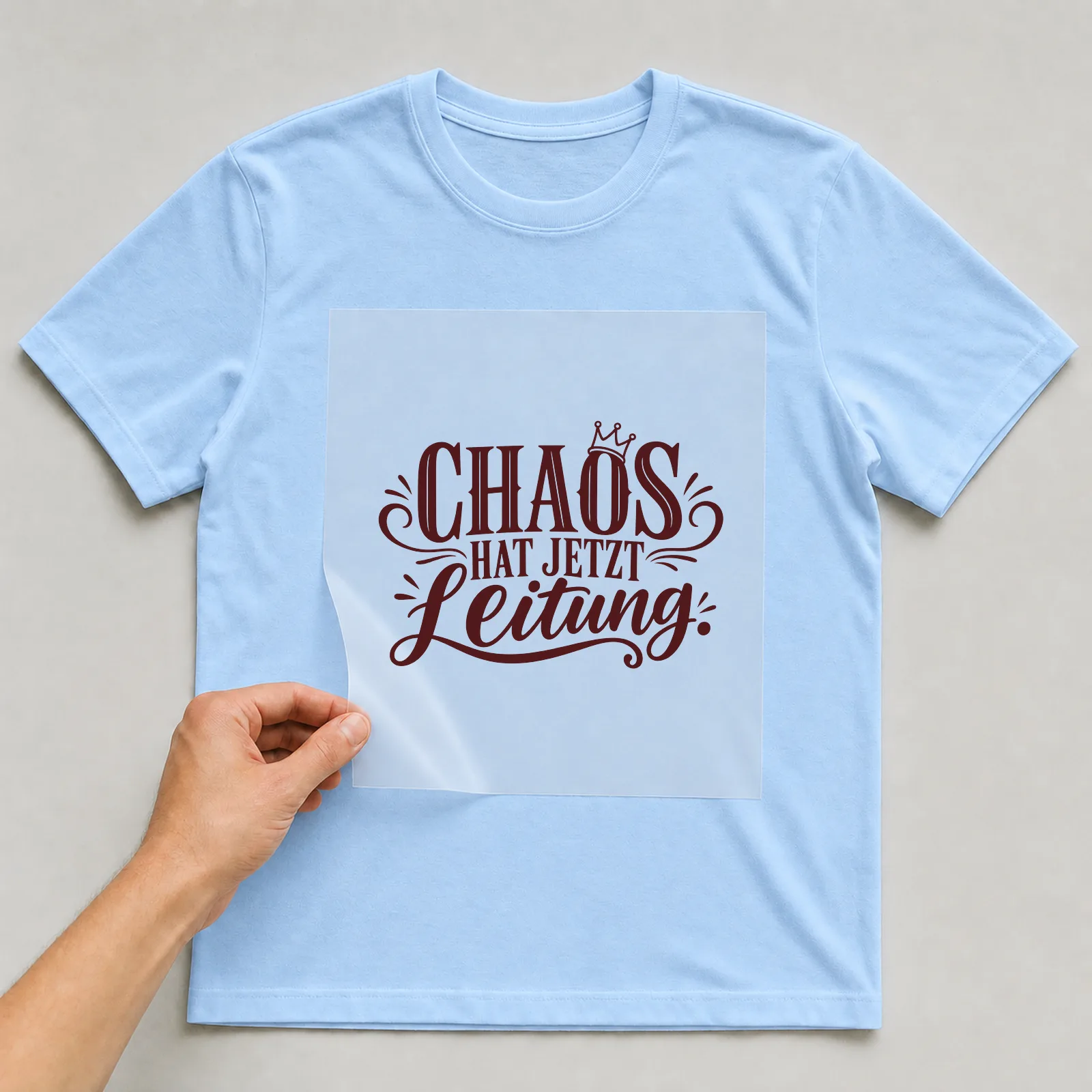 Bügelbild „Chaos hat jetzt Leitung“ auf hellblauem T-Shirt, Folie wird aufgelegt
