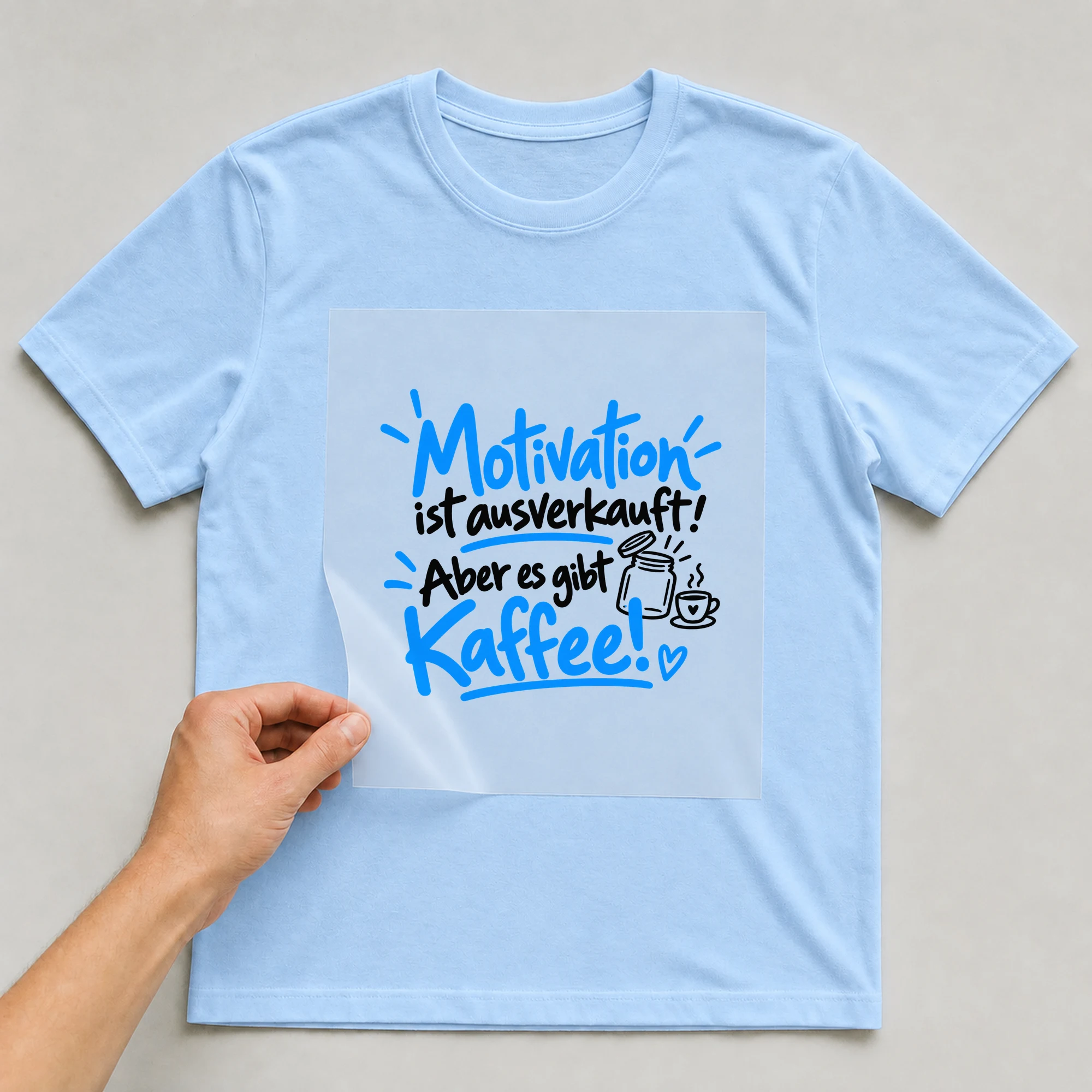 Bügelbild „Motivation ist ausverkauft! Aber es gibt Kaffee!“ auf babyblauem T-Shirt, Folie wird aufgelegt