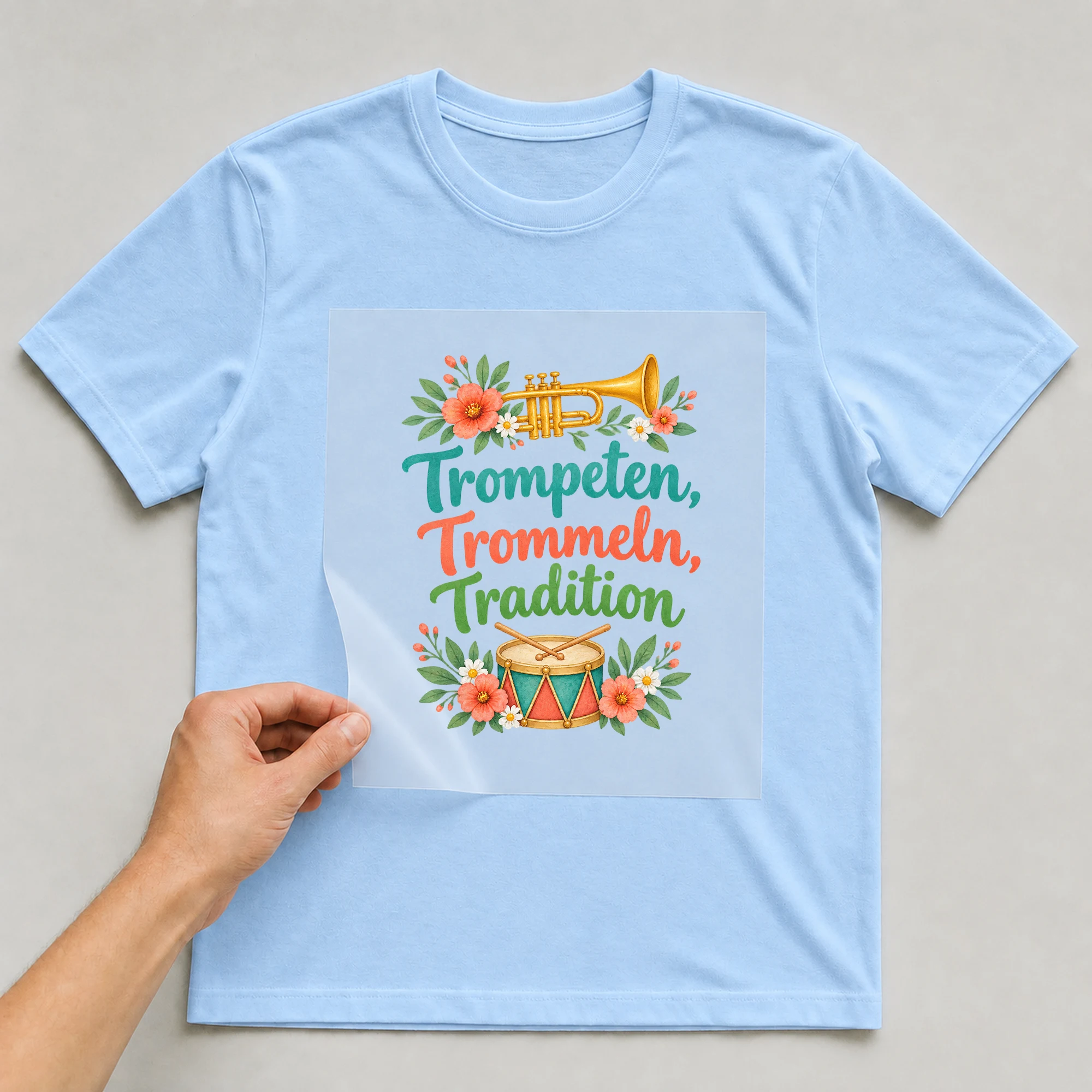 Bügelbild „Trompeten, Trommeln, Tradition“ auf babyblauem T-Shirt, Folie wird aufgelegt