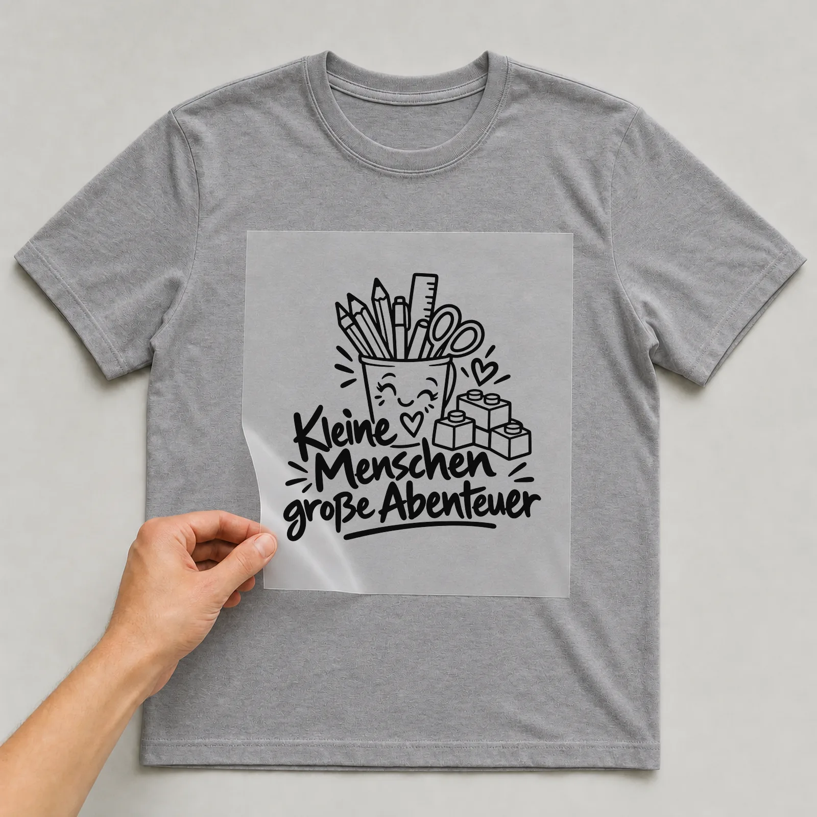 Bügelbild „Kleine Menschen große Abenteuer“ (klaren Konturen) auf grauem T-Shirt, Folie wird aufgelegt